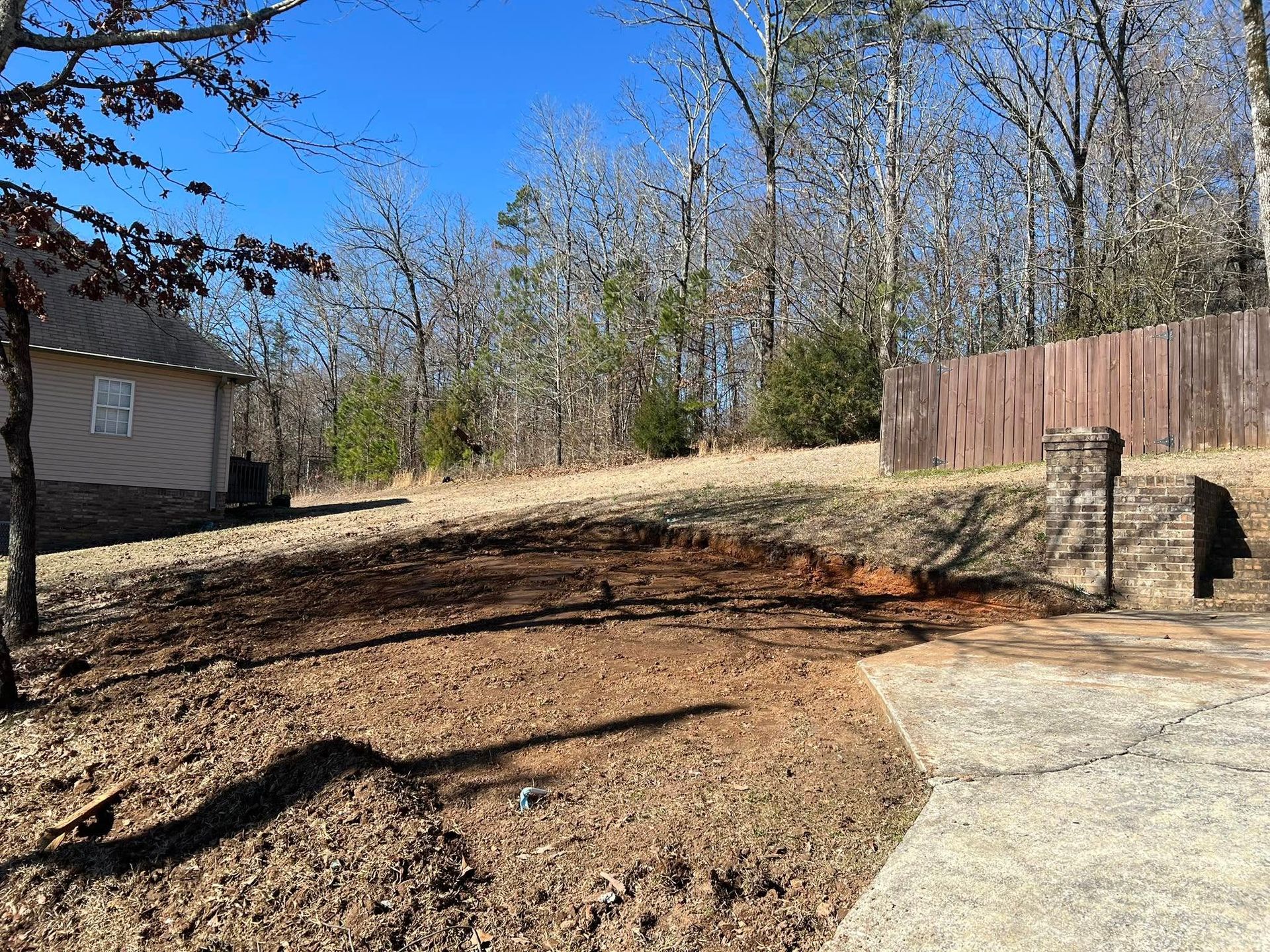 A cleared dirt plot beside a house, paved driveway, and stone pillar, framed by bare trees under a clear blue sky.