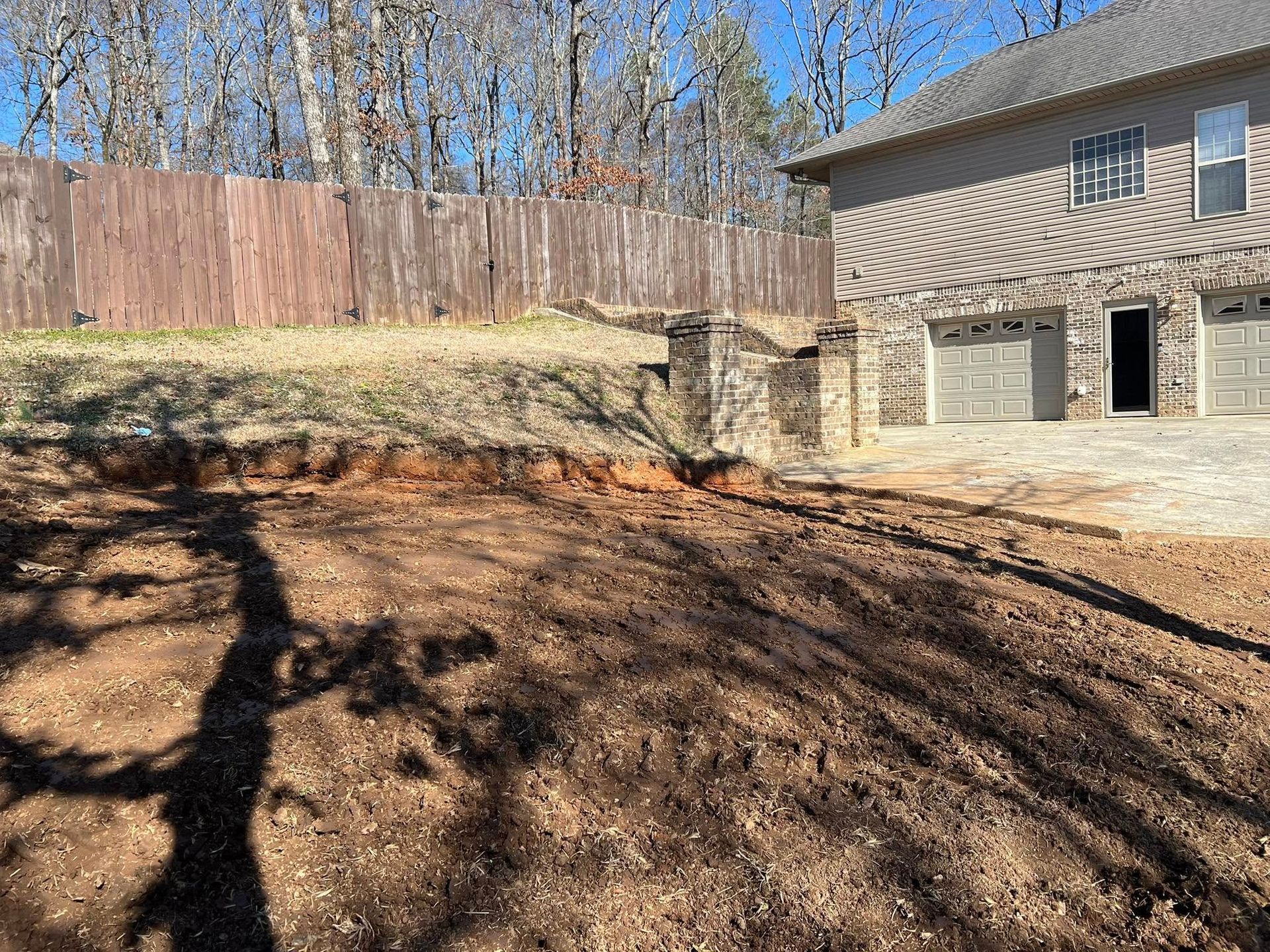 A sloping, recently cleared dirt yard leads up to a stone retaining wall and a house with a multi-car garage.