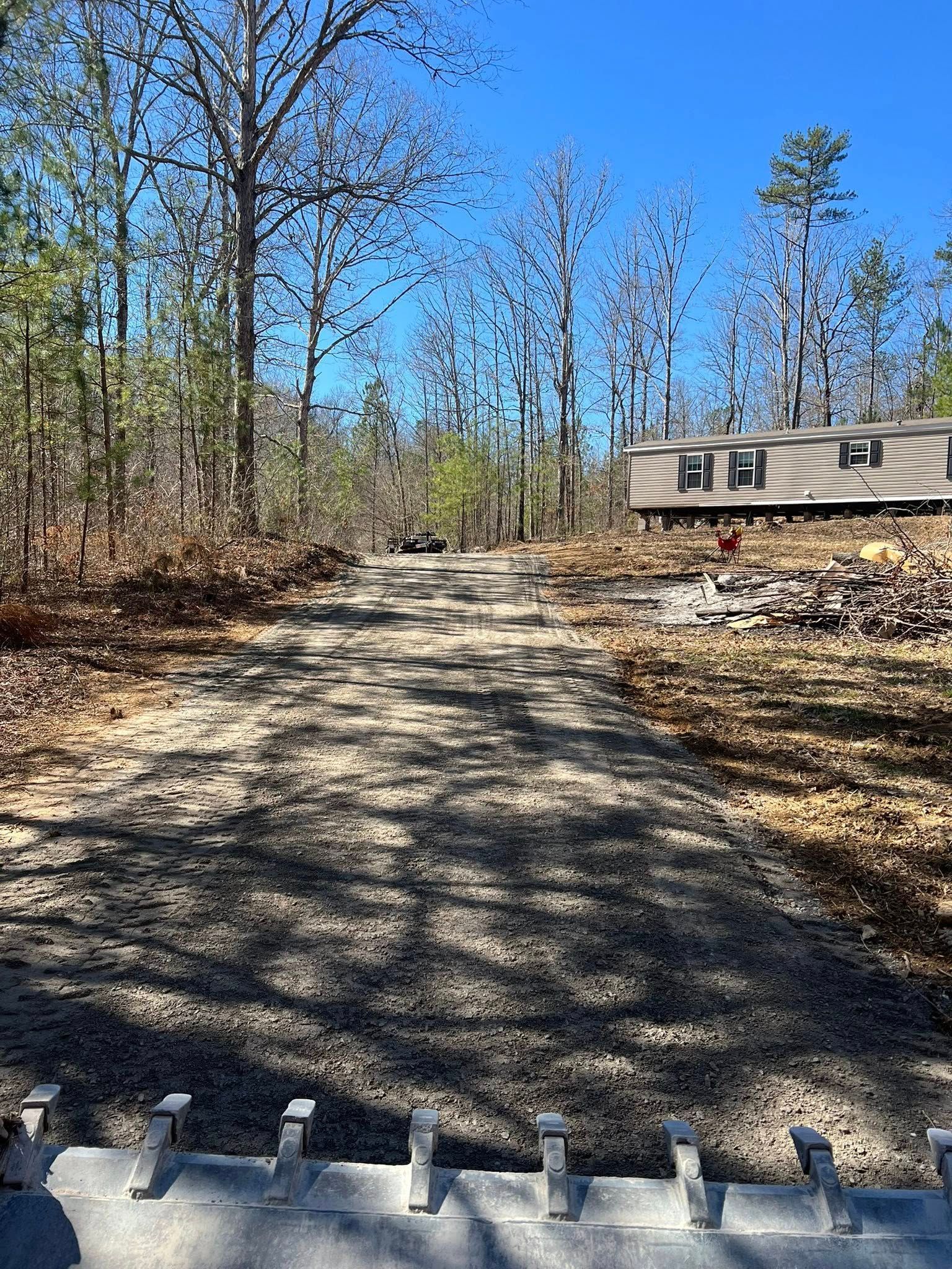 A gravel driveway leads upward through a wooded area toward a beige mobile home under a clear blue sky.