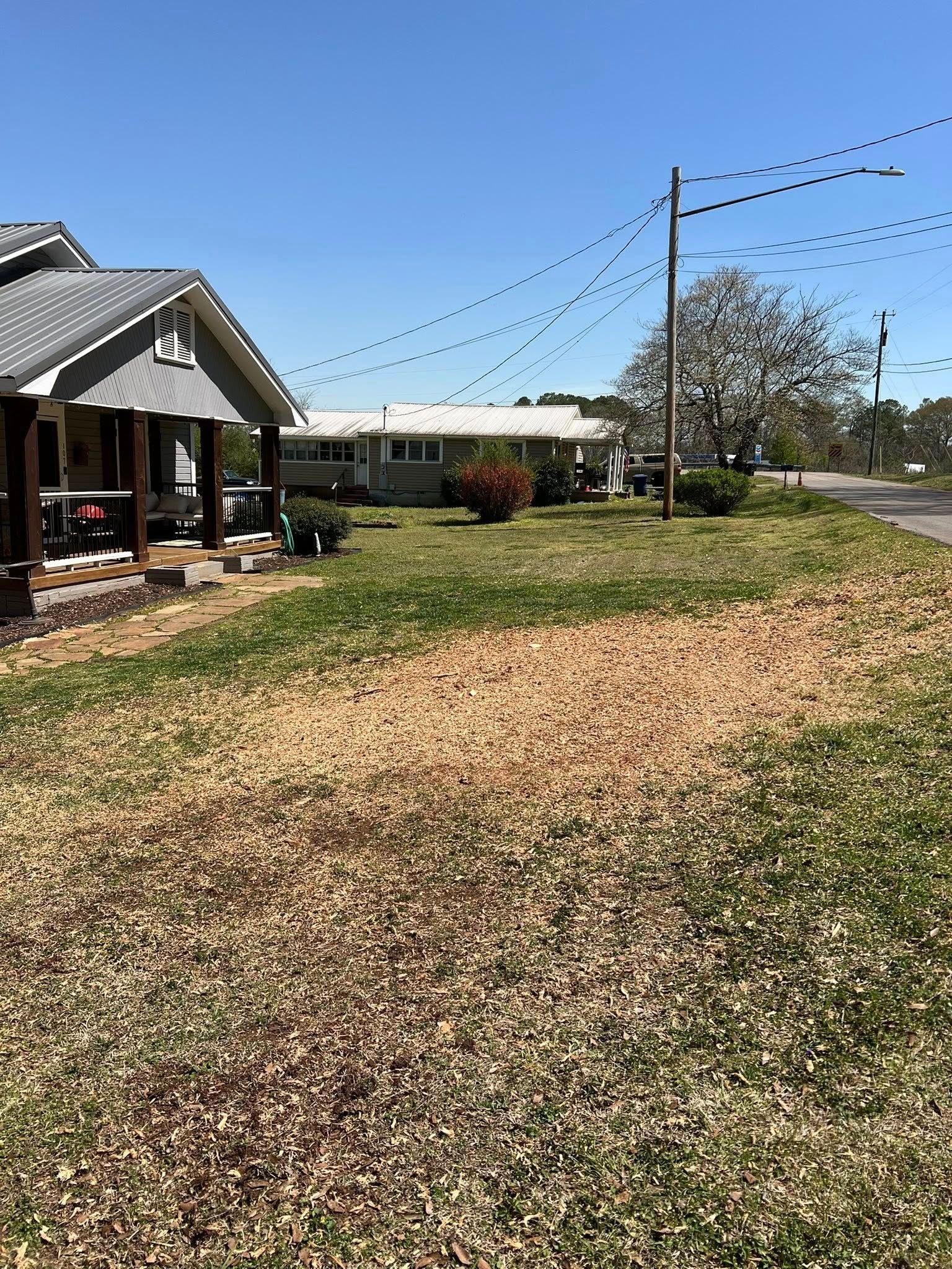 A house with a porch beside a grassy yard, with another house and a tree in the distance under a clear blue sky.