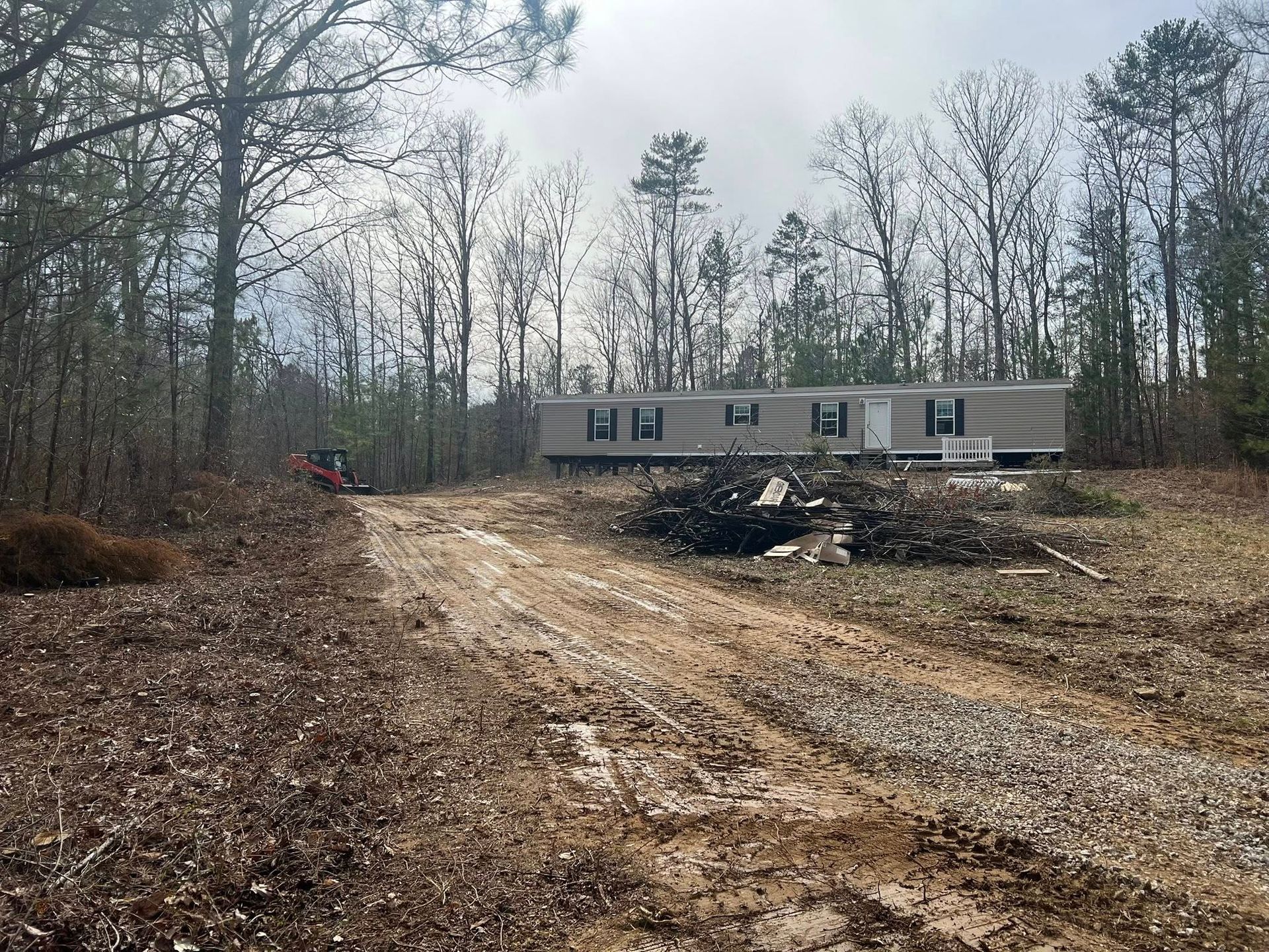 A beige mobile home sits atop a grassy clearing surrounded by a dense forest, reached by a long, gravel driveway.