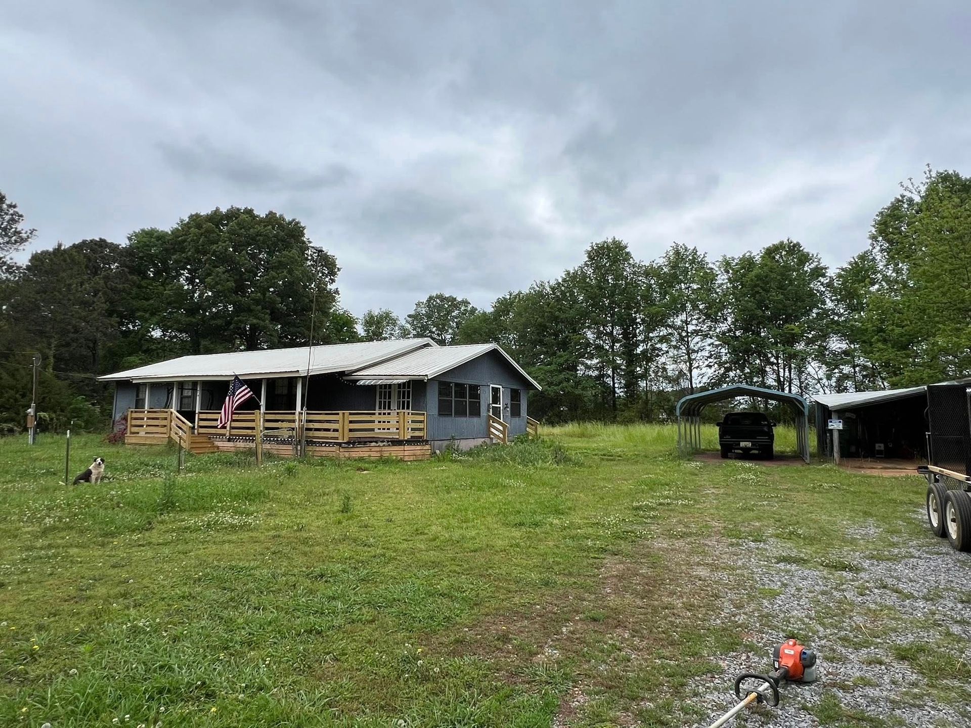 A gray, single-story house with a wooden porch and American flag, near a gravel driveway with a carport and vehicle.