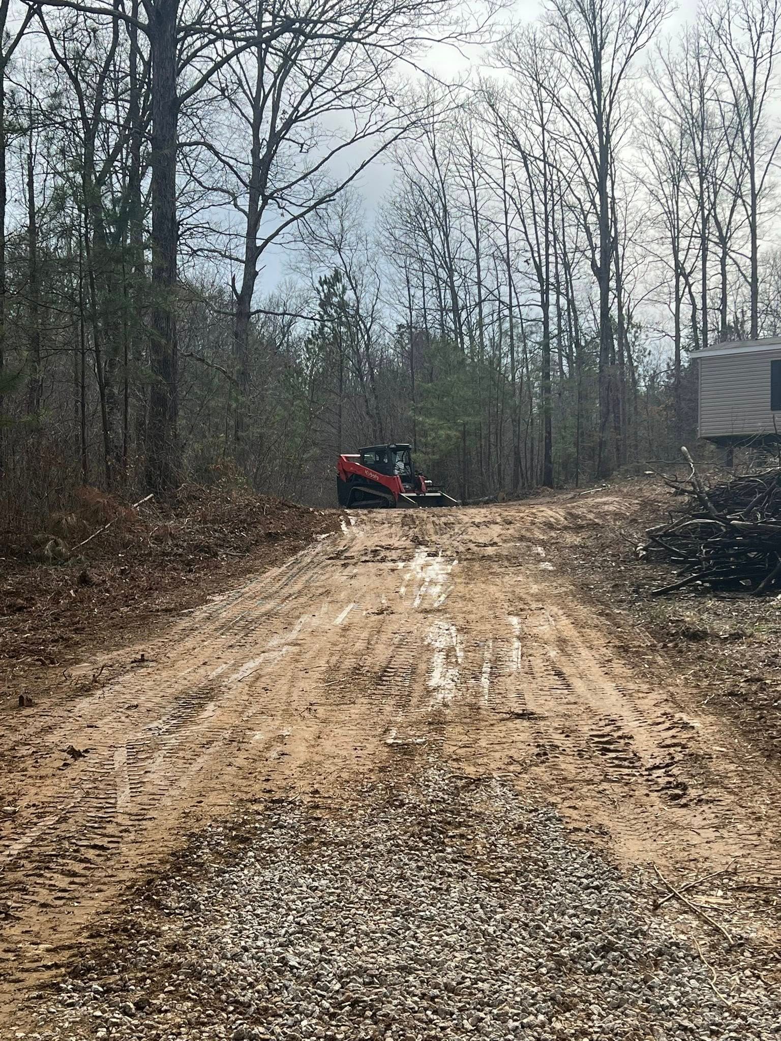 A red skid steer sits on a dirt path in a wooded area, with a gravel surface in the foreground.