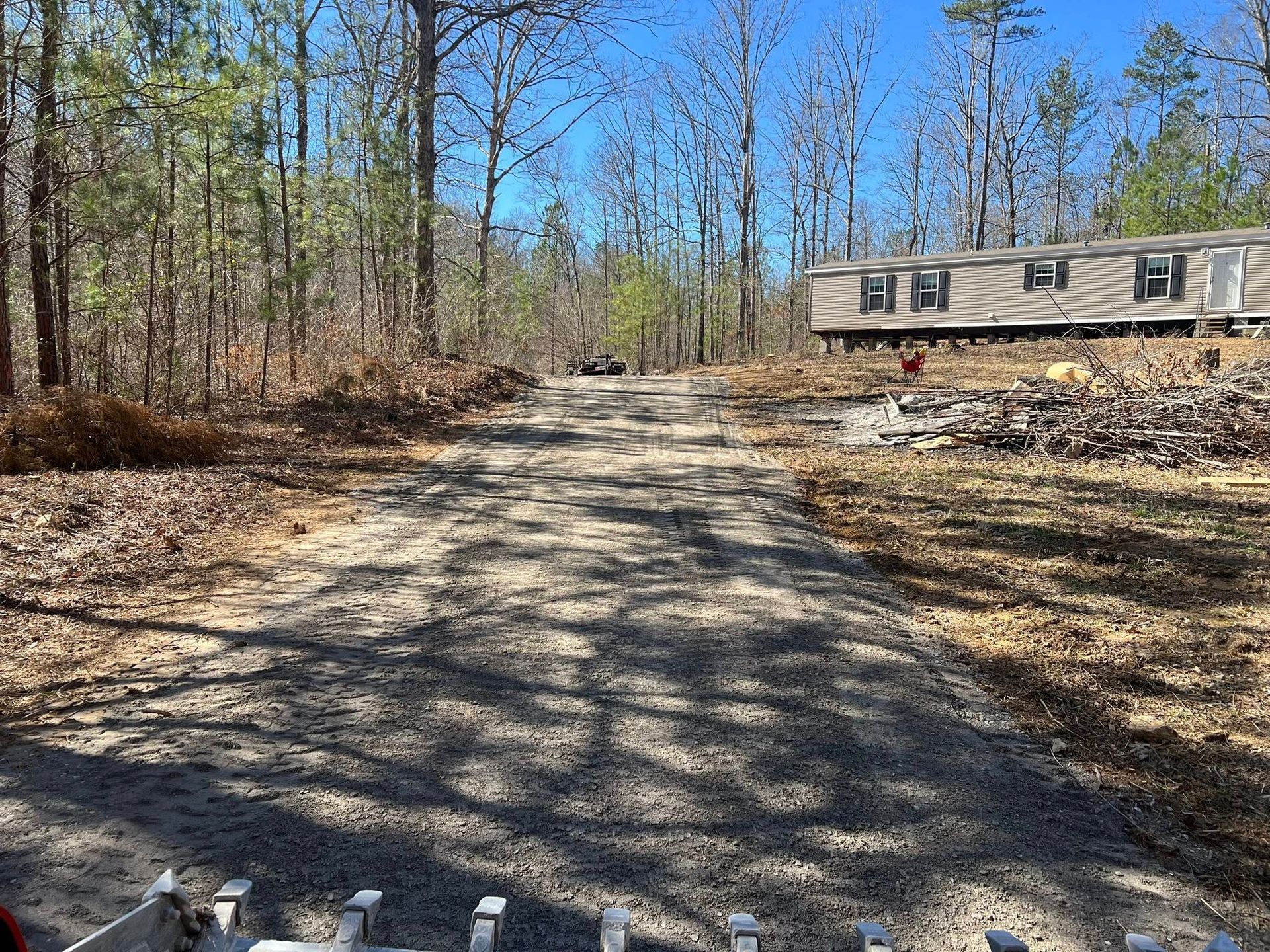 A gravel driveway leads through a wooded area toward a light-colored mobile home on a sunny day.