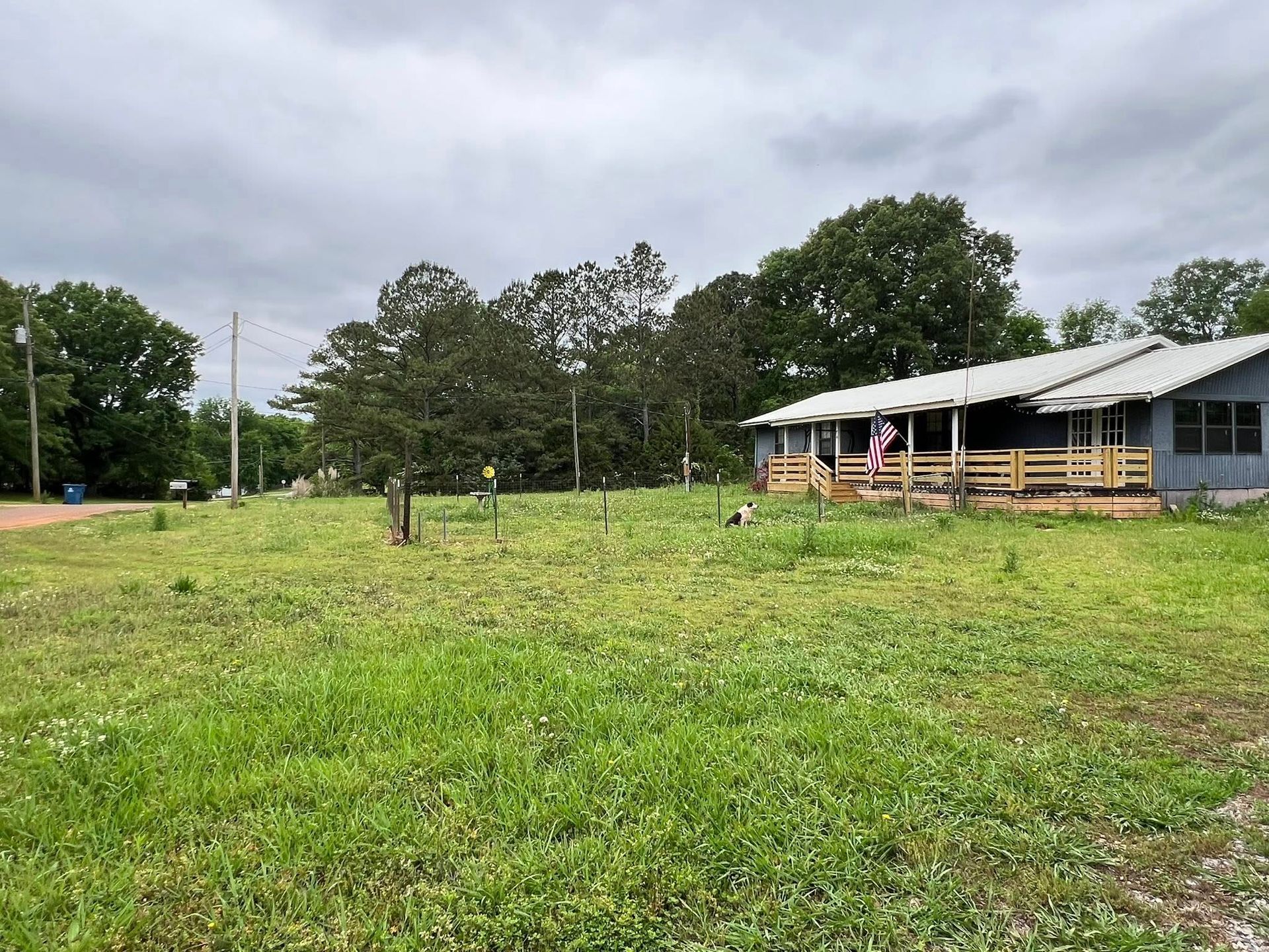 A gray house with a wooden porch sits on a grassy lot under a cloudy sky, with trees in the background.