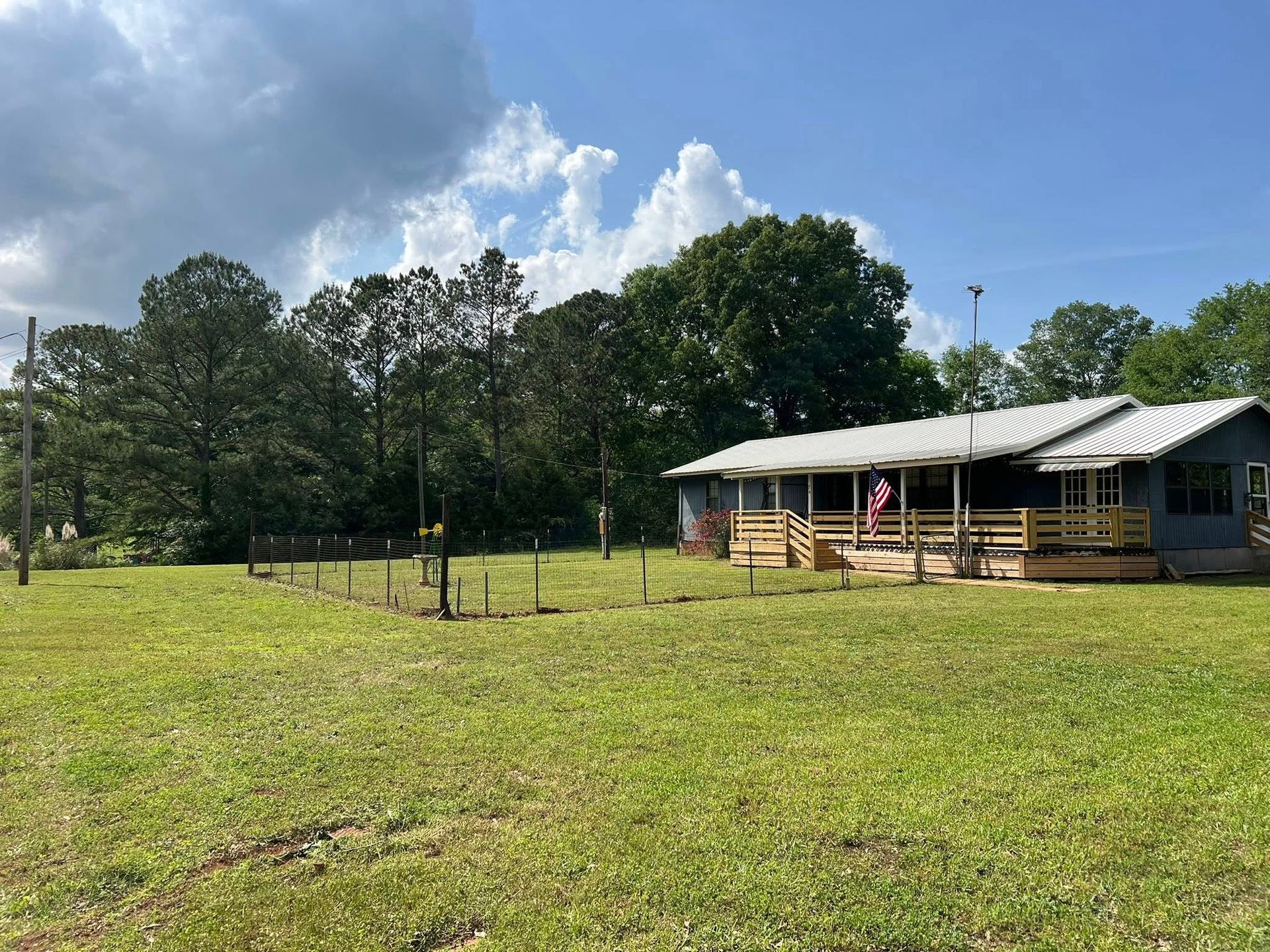 A gray house with a wide wooden porch and an American flag sits in a sunny, grassy yard surrounded by mature trees.