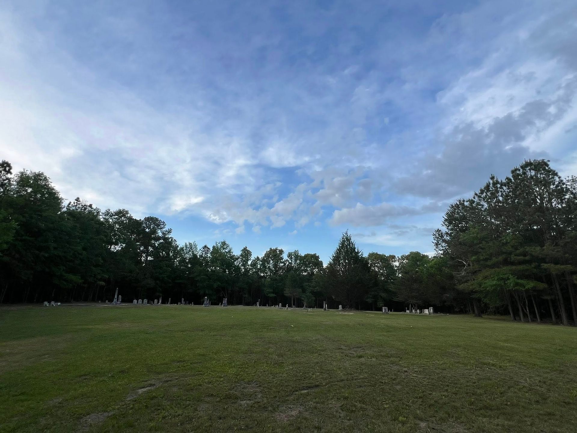 A grassy field slopes upward toward a line of trees under a bright blue sky with wispy clouds, with gravestones visible.