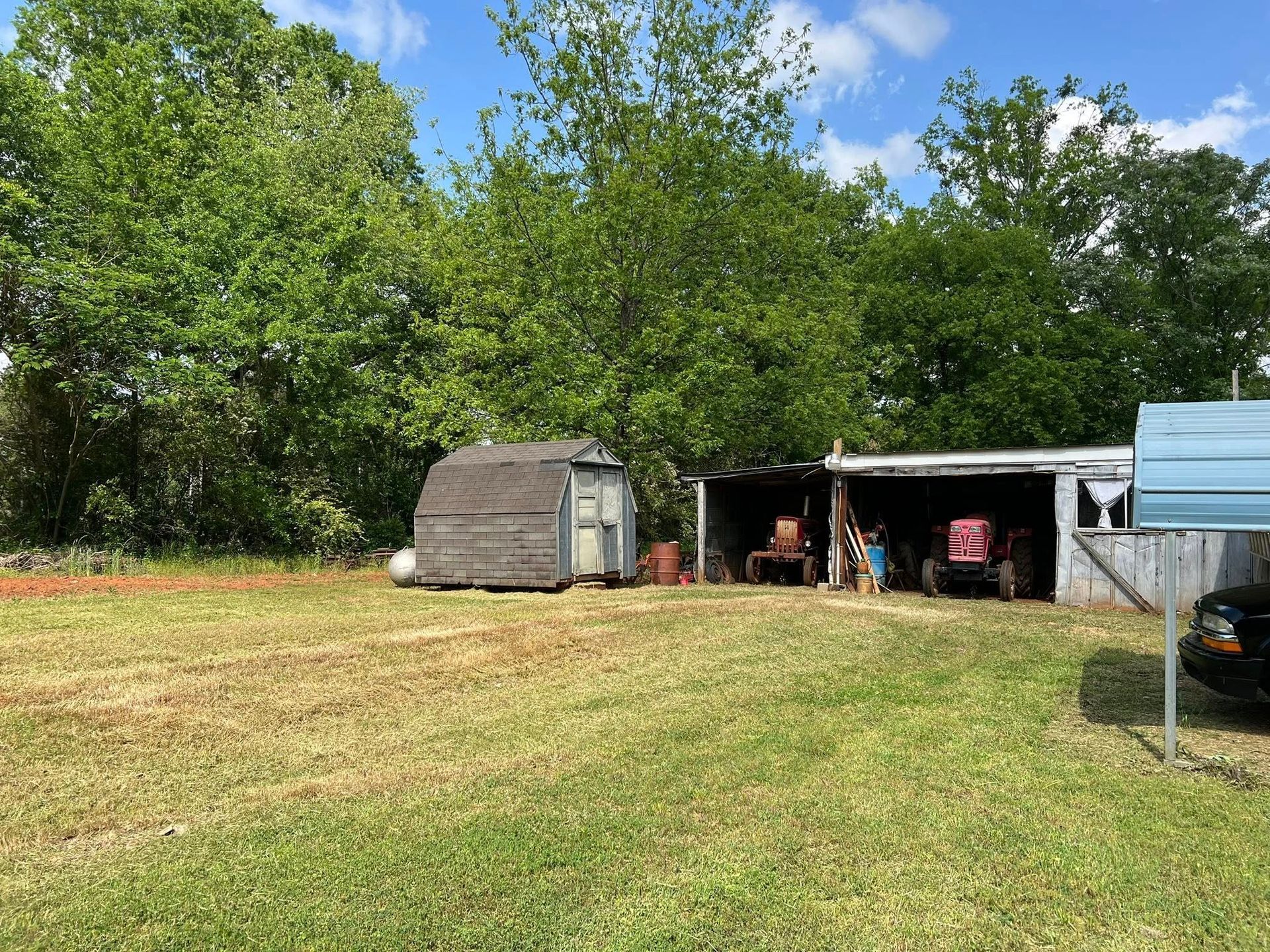 A small, weathered shed sits next to a carport sheltering two red tractors on a grassy lot under a bright, sunny sky.