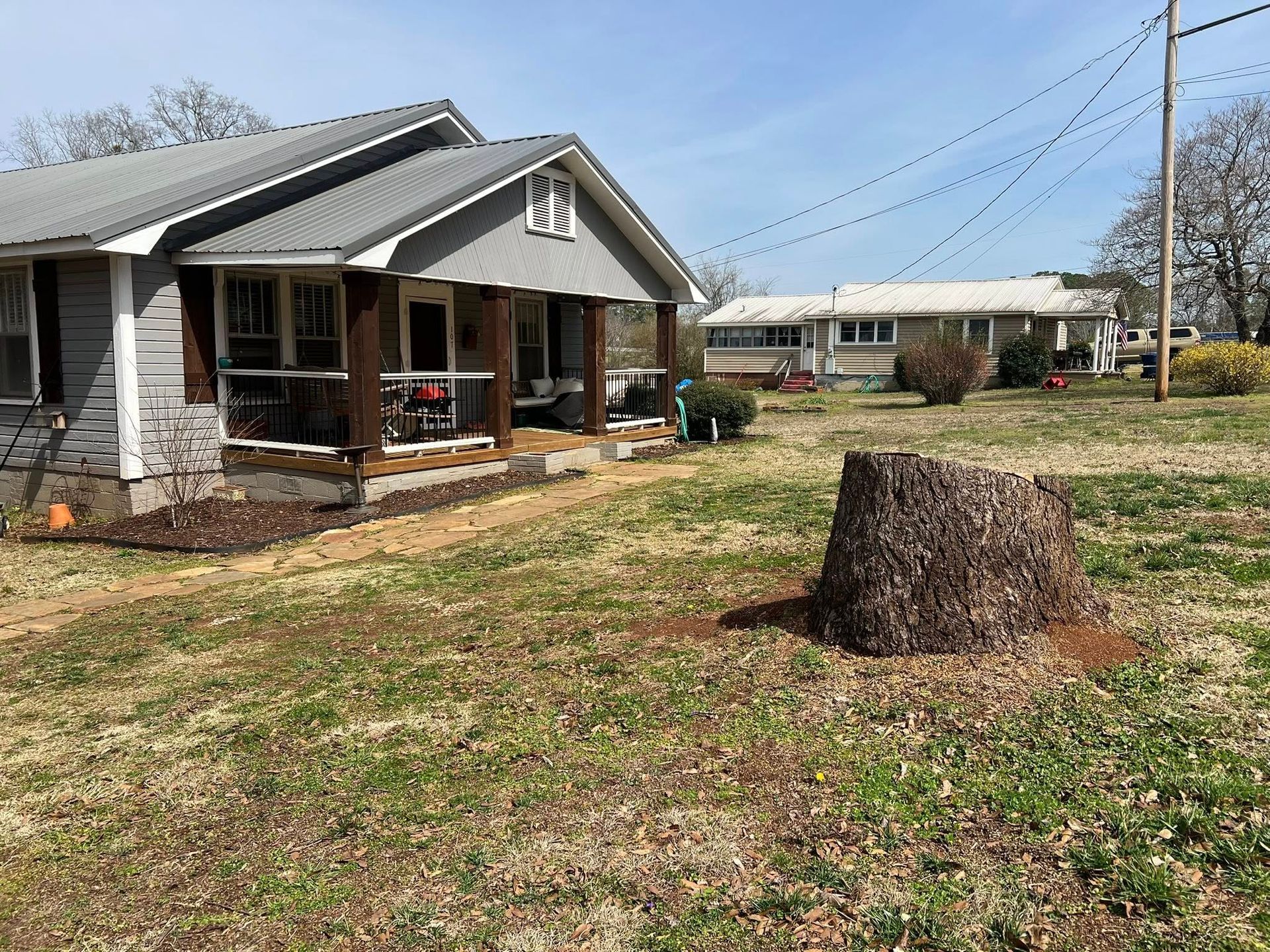 A grey, single-story house with a front porch sits on a grassy lot next to a large tree stump, with another home nearby.