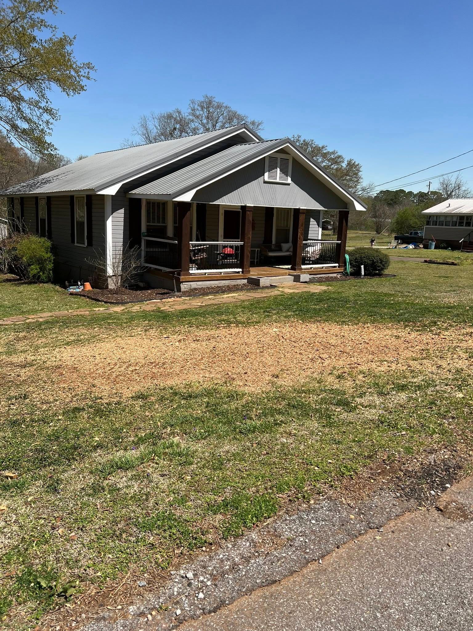 A small, gray-sided house with a front porch and a gabled roof sits on a grassy lot under a clear blue sky.