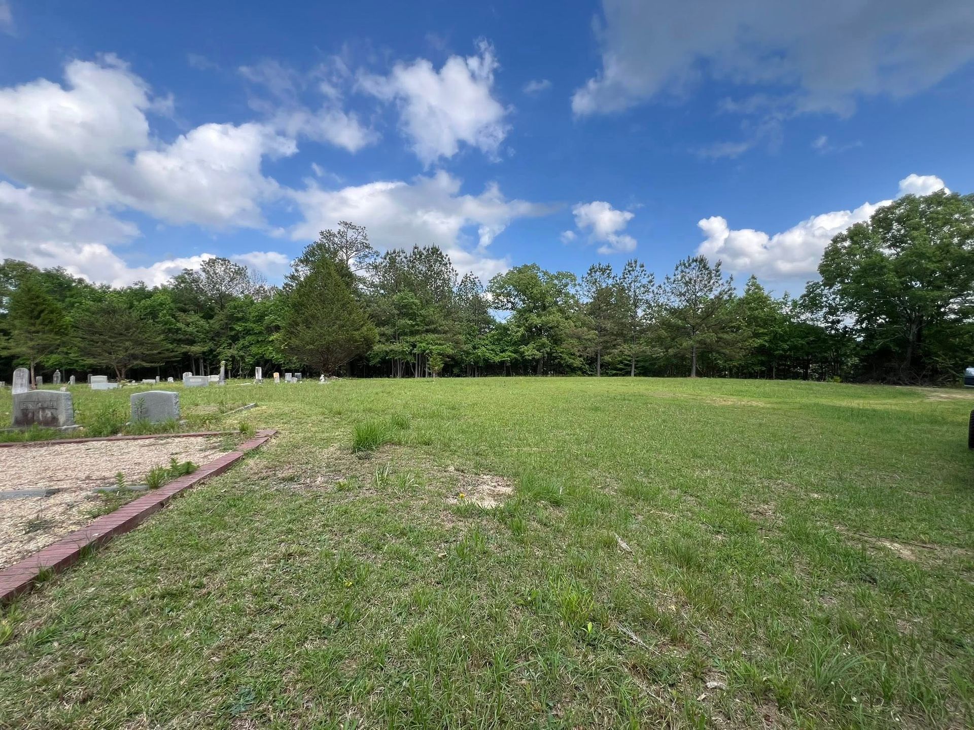 A grassy cemetery under a bright blue sky with scattered clouds, featuring gravestones near a stone-bordered pathway.