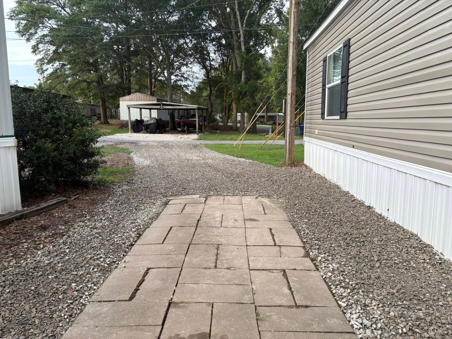 A stone walkway leading through a gravel driveway toward a metal carport next to a house with beige siding.