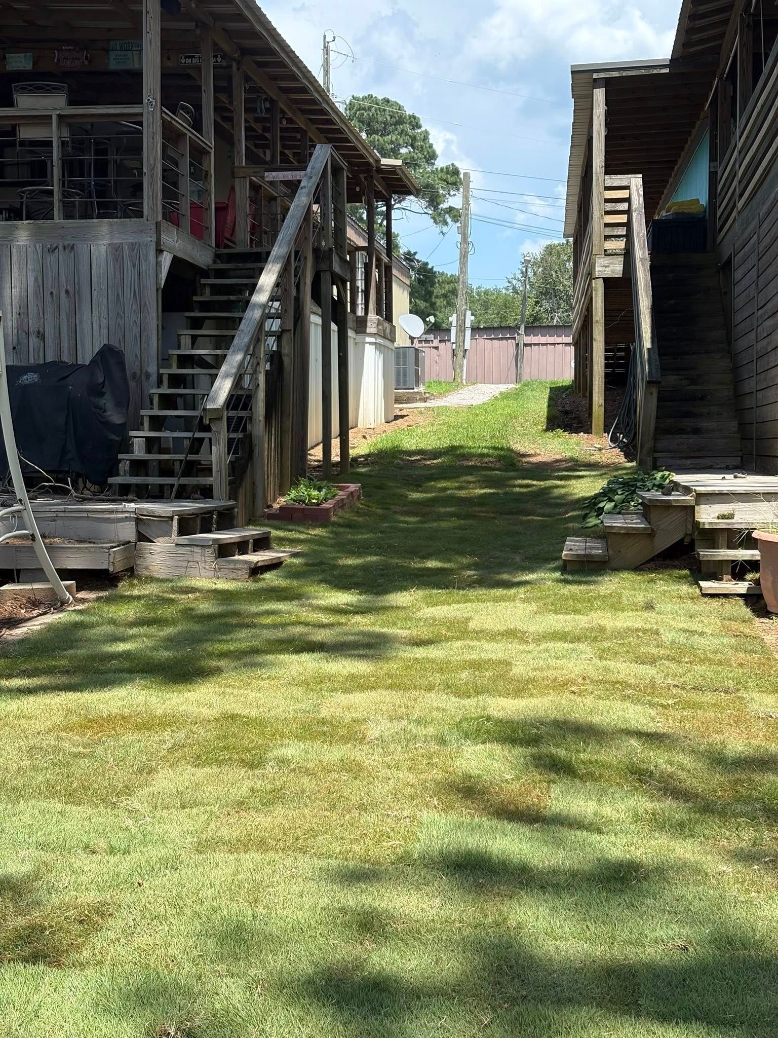 A grassy path between two elevated wooden buildings with outdoor stairs, under a clear, sunny sky.