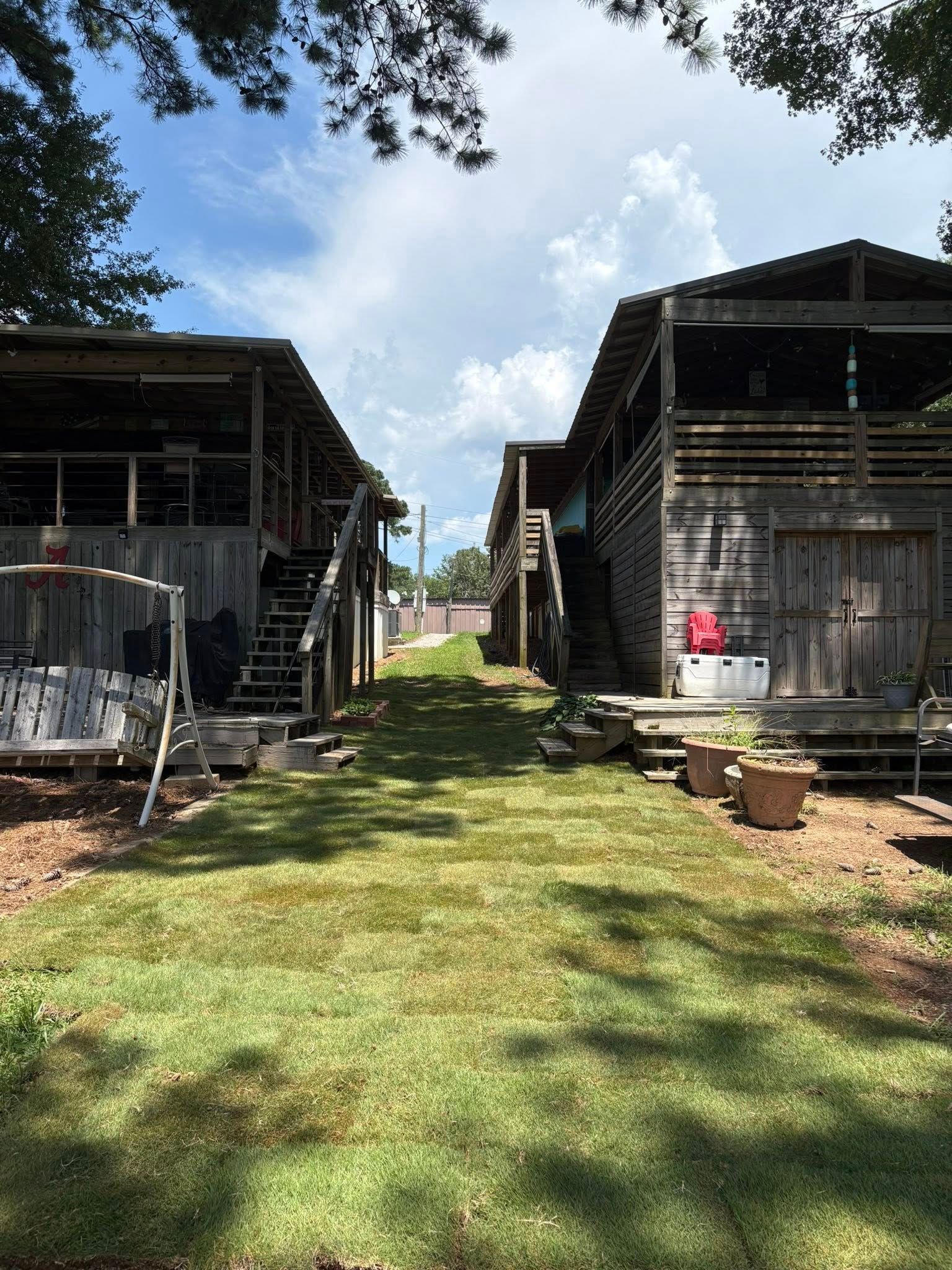 A grassy path between two rustic, elevated wooden cabins under a bright sky with trees above.