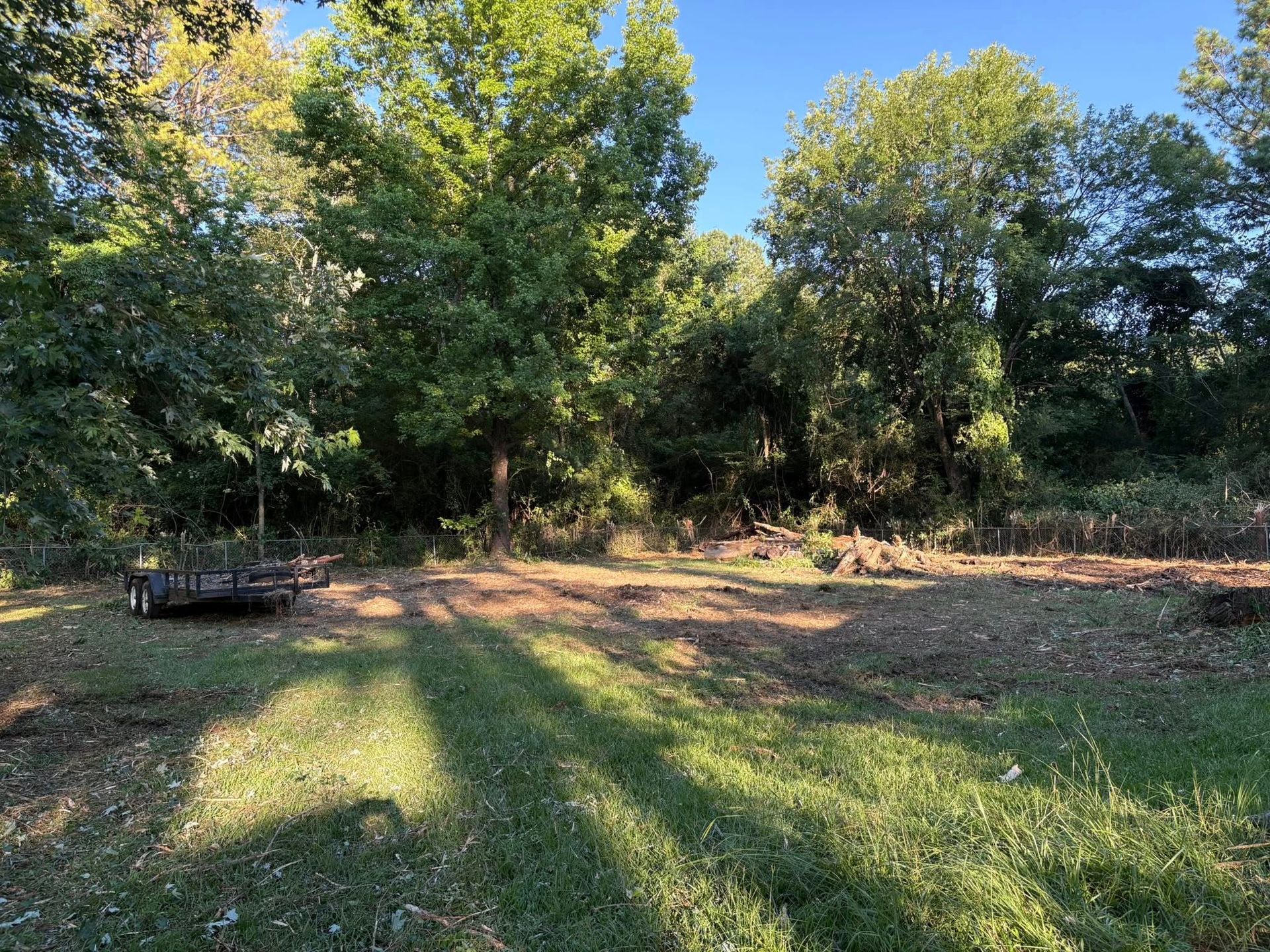 A cleared dirt lot in a rural, wooded setting with a small black utility trailer parked on the left.