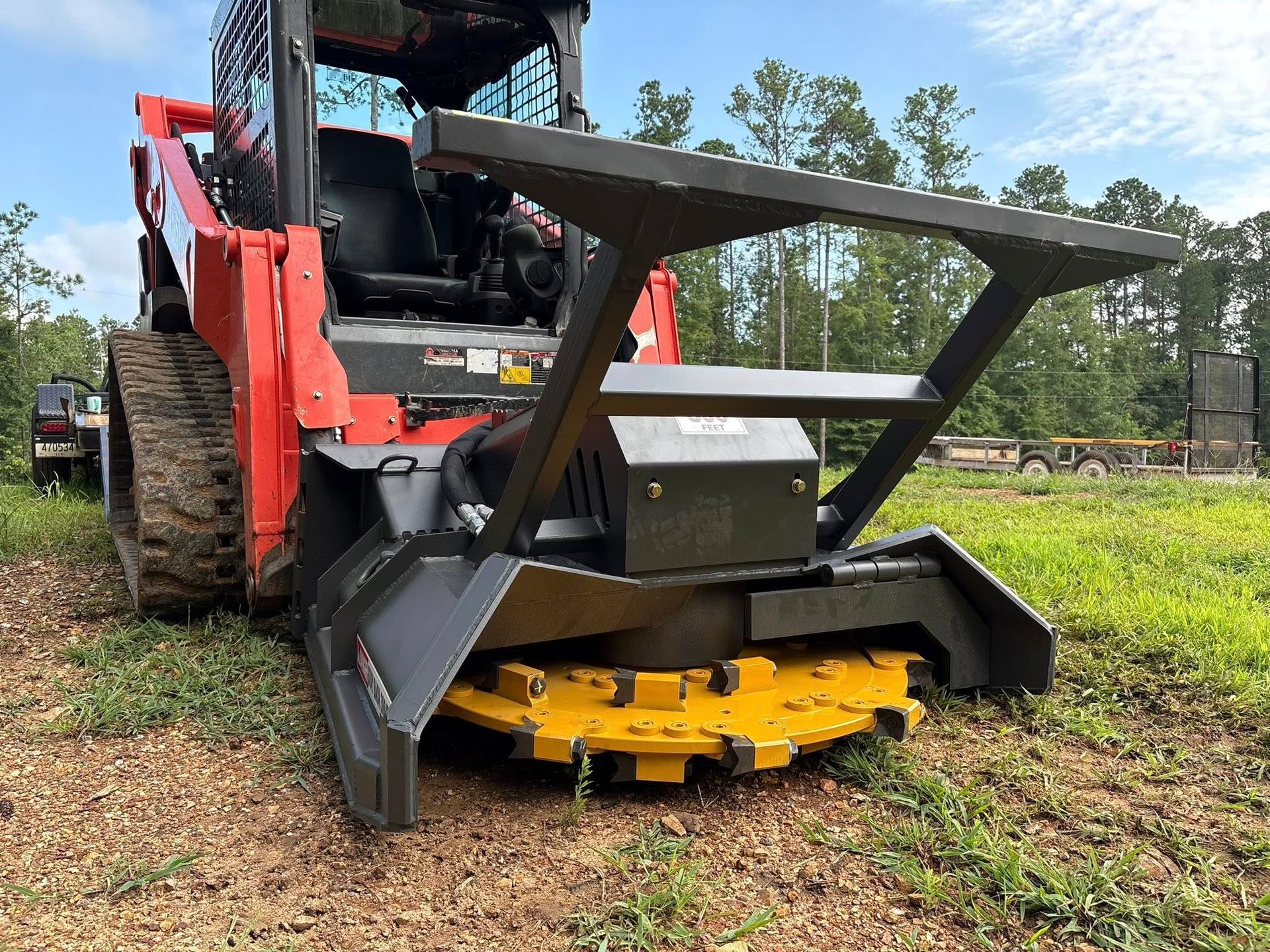 An orange skid steer loader with a yellow rotary brush cutter attachment sits on a grassy field.