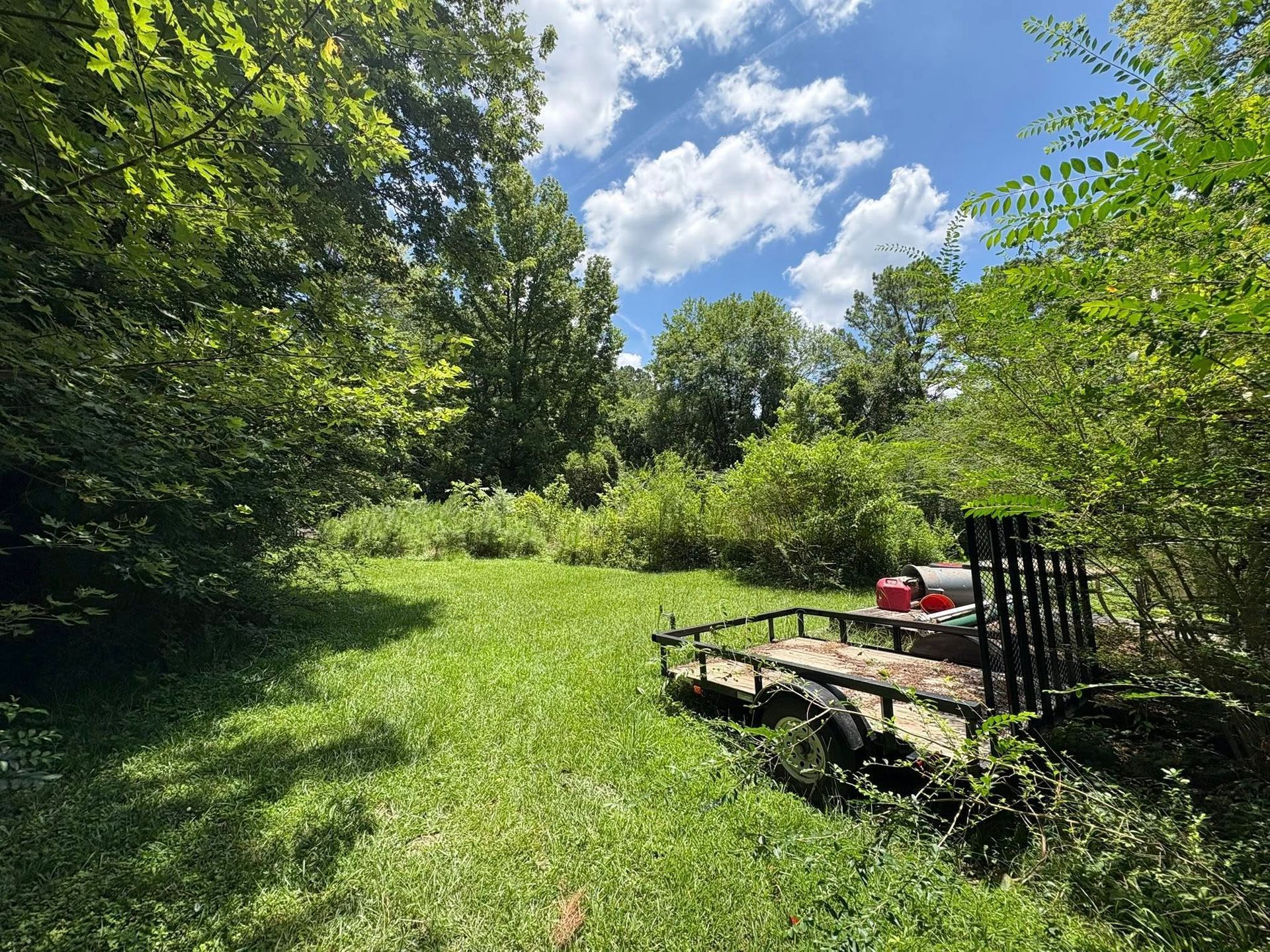 A flatbed utility trailer sits in a lush green, tree-lined meadow under a blue sky with scattered clouds.