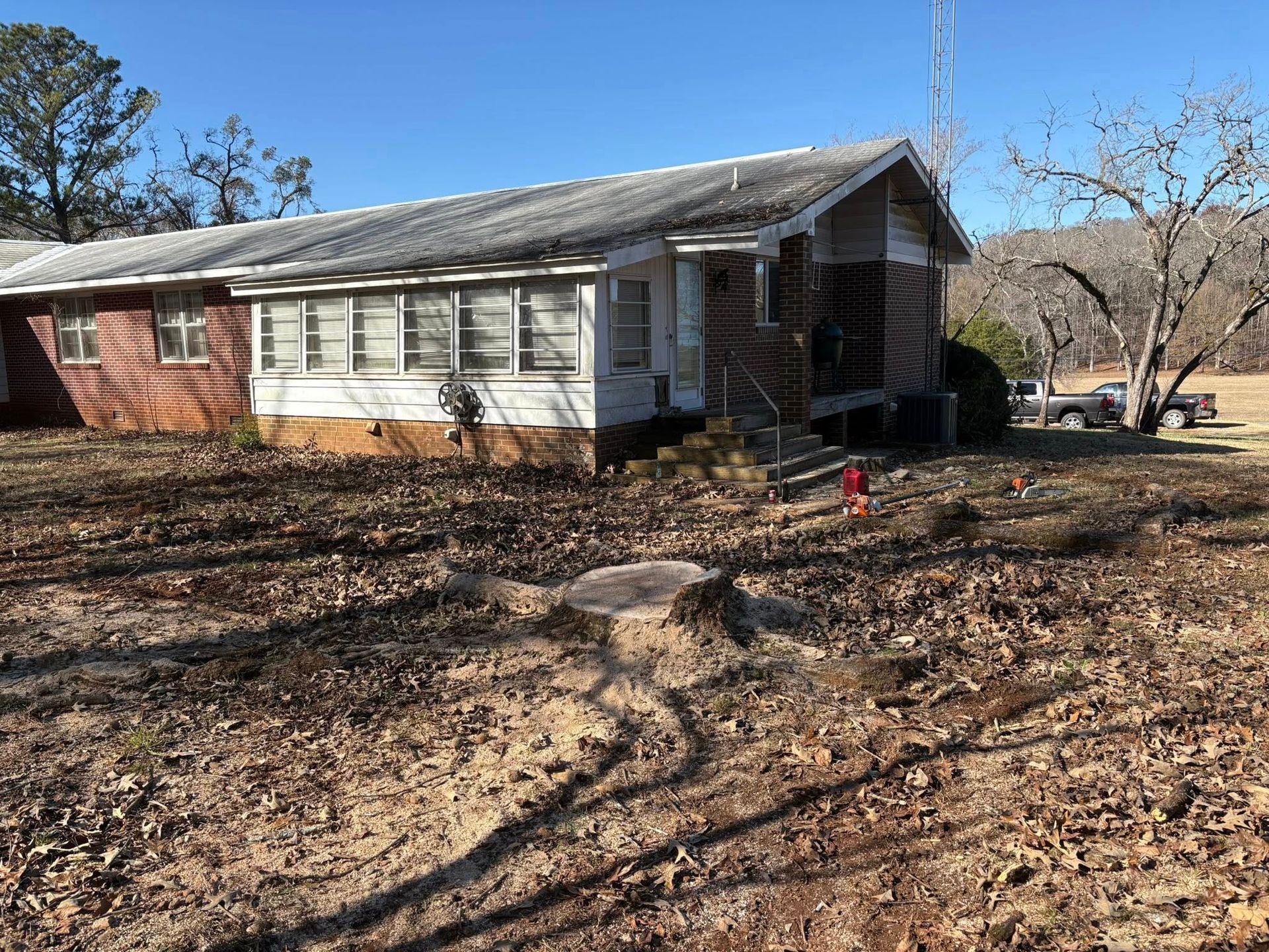 A single-story, white-sided house with a low-pitched roof sits in a yard covered in fallen leaves and a tree stump.