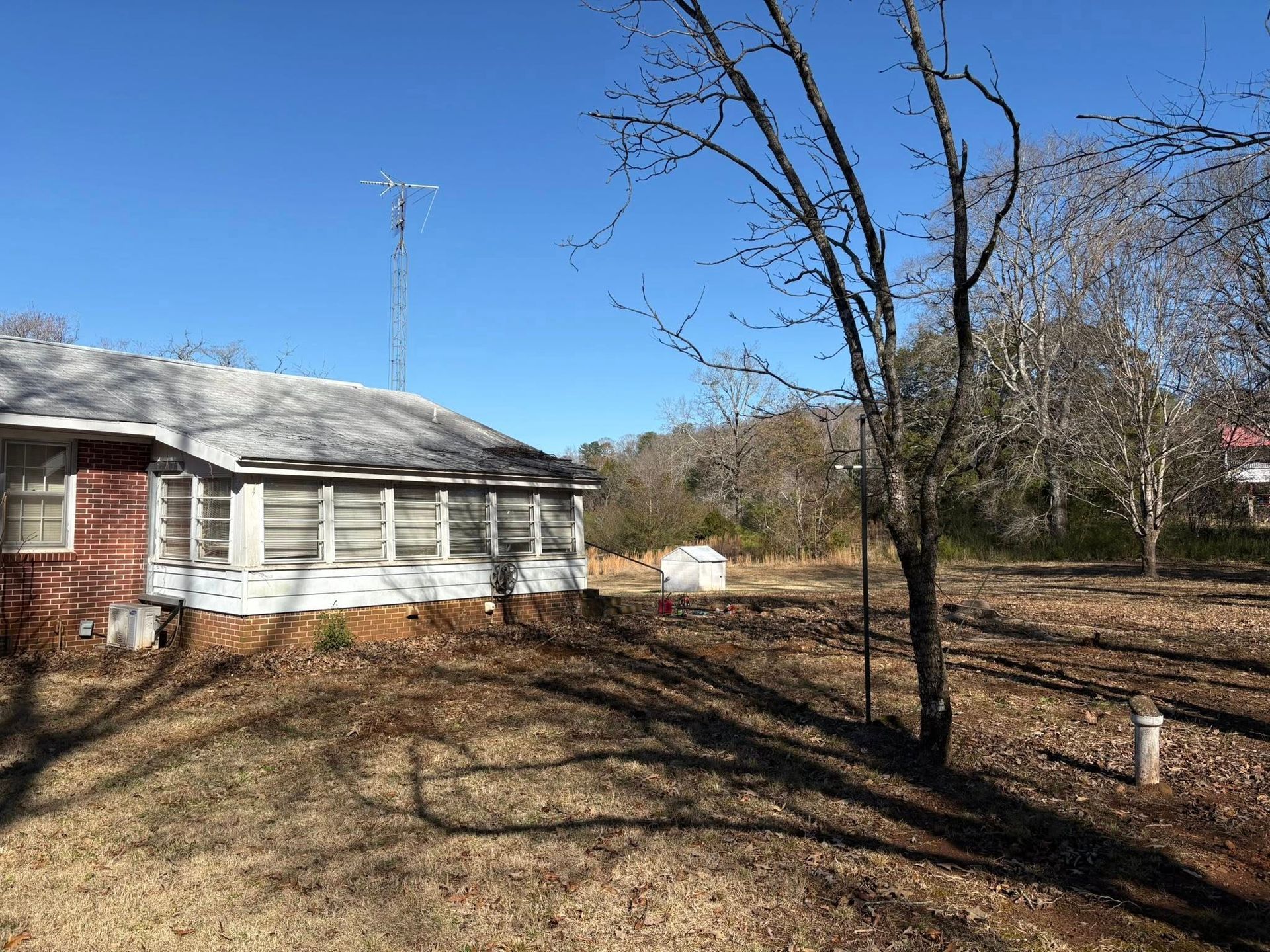 A side view of a house with a white enclosed porch under a clear blue sky, surrounded by leafless trees and a dry yard.