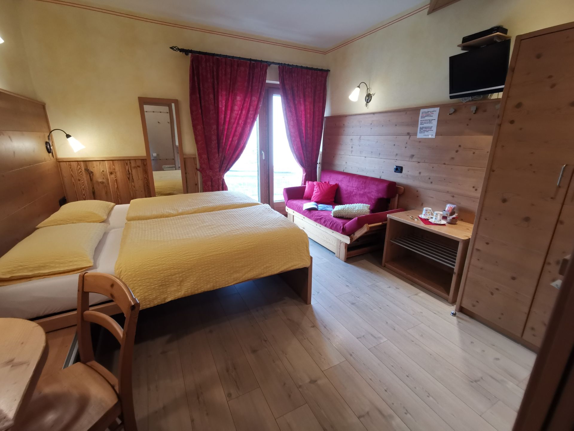 Accogliente camera d'albergo con letto matrimoniale, divano, mobili in legno, tende rosse e balcone.