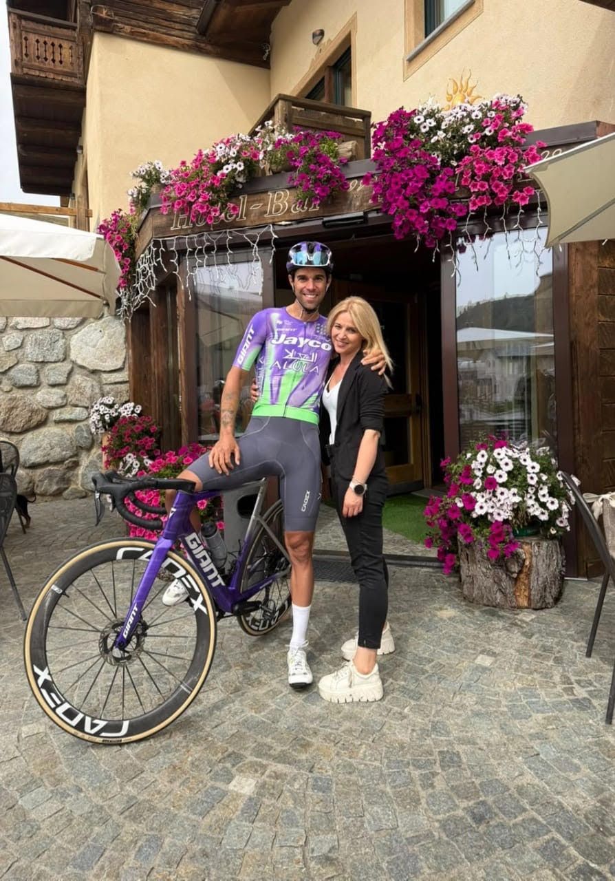 Ciclista con la bicicletta in piedi accanto a un cartello di fronte a un hotel di montagna. Montagne sullo sfondo.