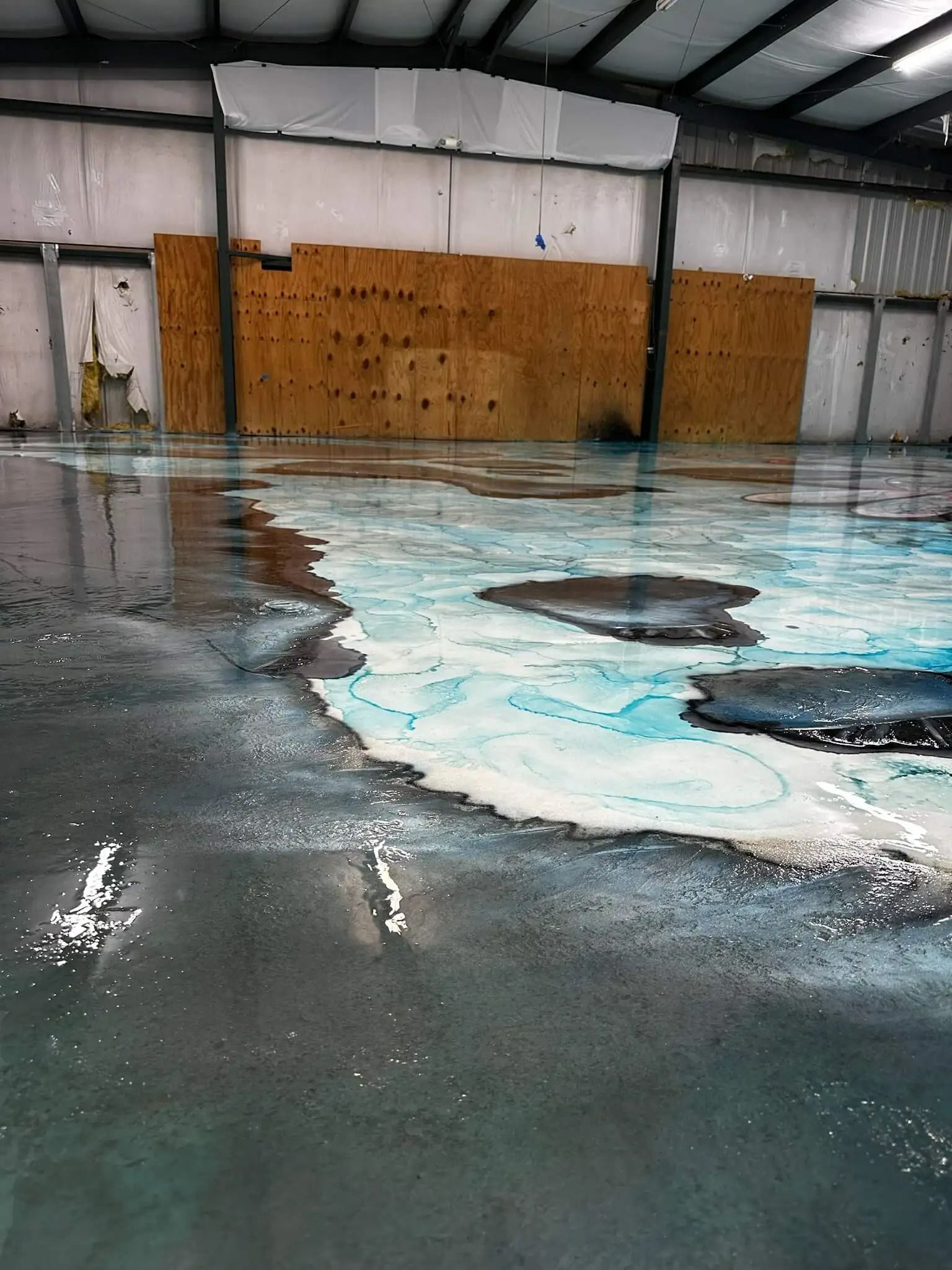 A flooded warehouse floor reflecting the ceiling and wall; blue, white, and grey paint and water cover the concrete.