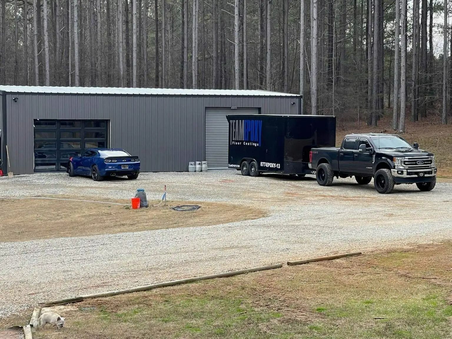 Dark gray truck pulling a trailer in front of a metal building with a blue sports car parked outside.