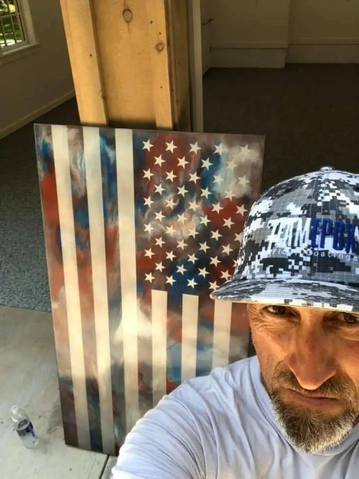 Man in a white shirt and digital camo hat poses next to an American flag art piece. The artwork features red, white, and blue colors.