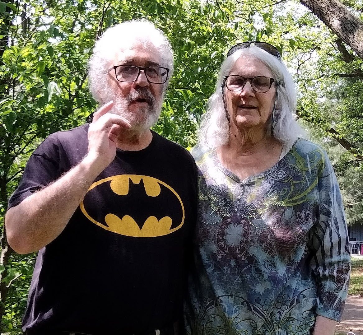 Older white straight couple in front of trees. The man has short curly white hair and square glasses and the woman has long curly hair.