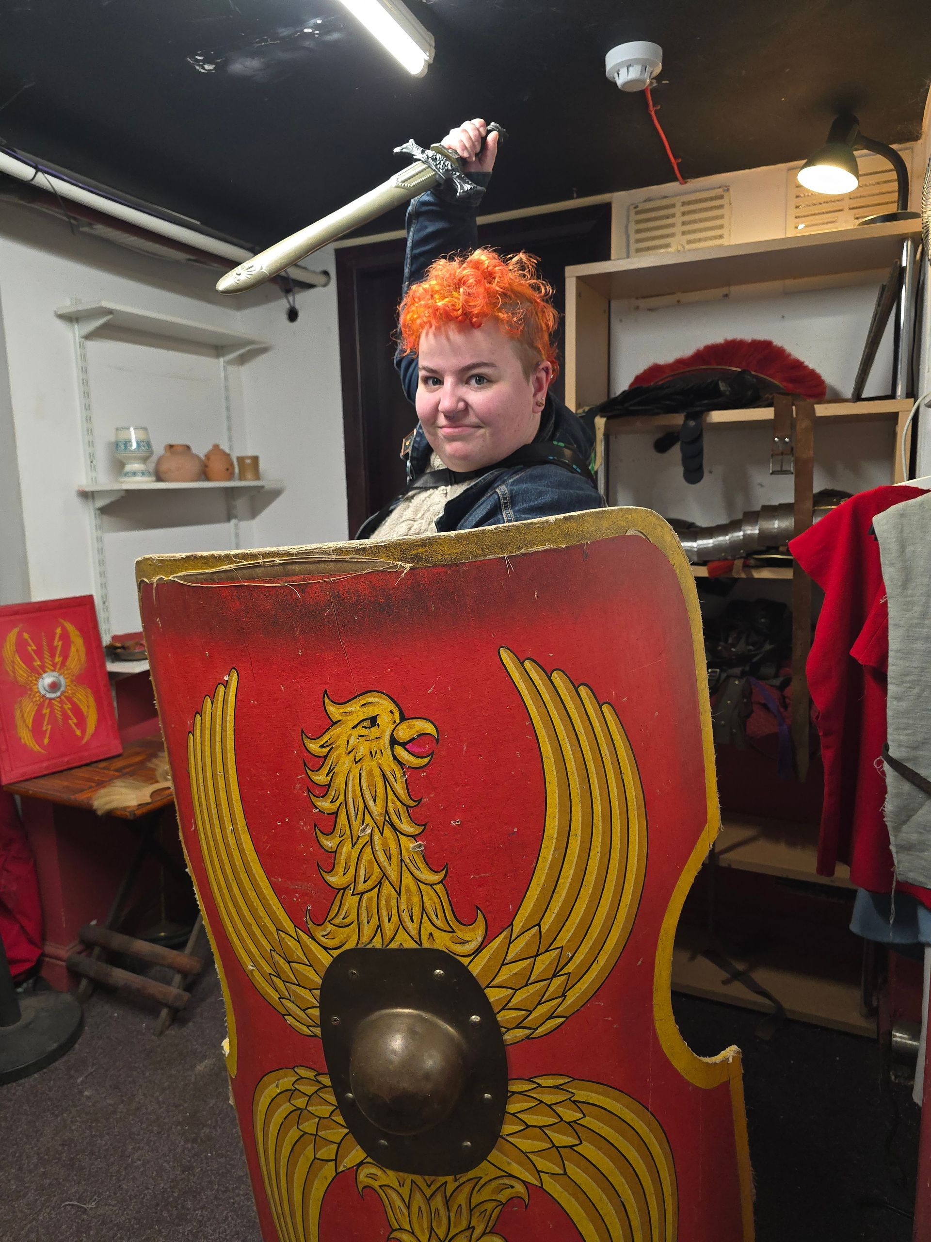 A white person with a red-orange mohawk holding a roman shield weilding a sword