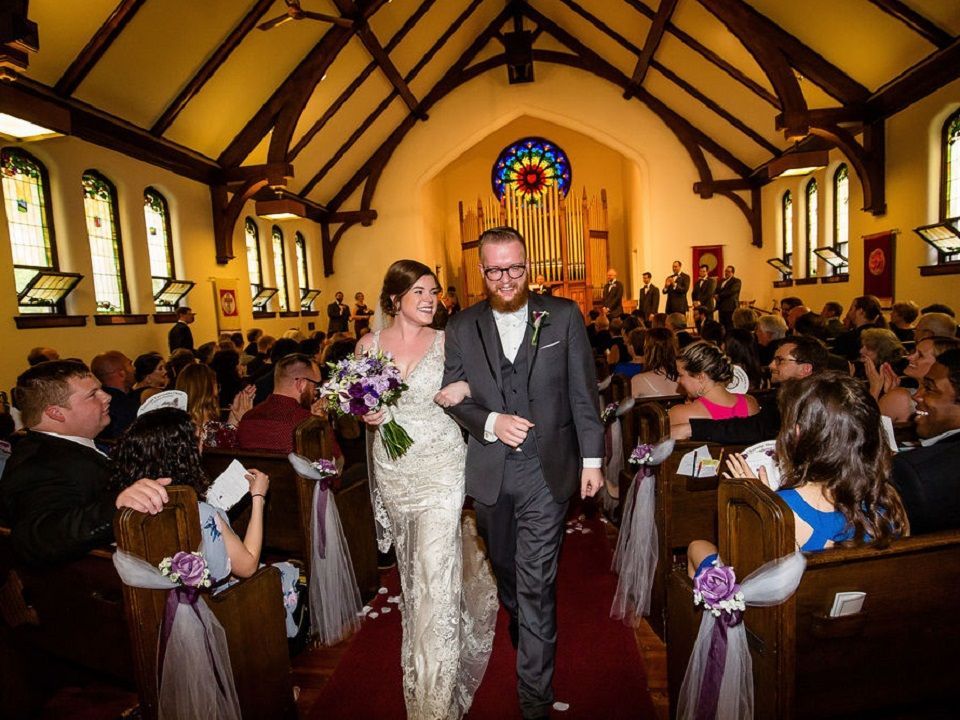 a white couple wearing a wedding dress and a black suit walk down the sanctuary aisle full of guests