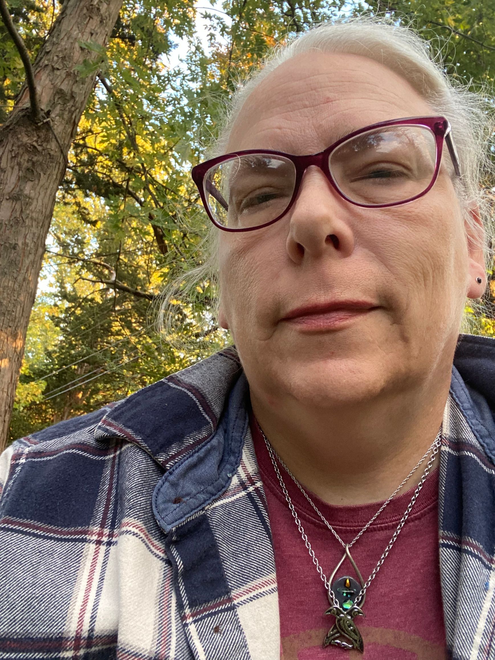 A white woman with gray hair and purple rimmed glasses smiling in front of some trees