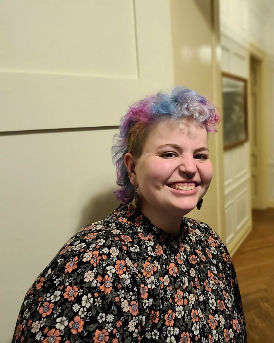 Alyssa Knights has a curly dyed mullet. Their hair is blue, pink and purple. They are wearing large leaf earrings. 