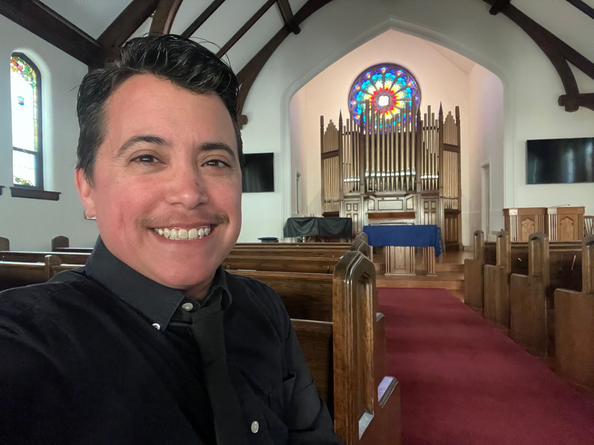 Nonbinary hispanic person with black short hair in the sanctuary with the rose window behind them