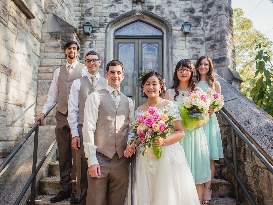 A group of people wearing their wedding best stand outside of the stone church building with flowers