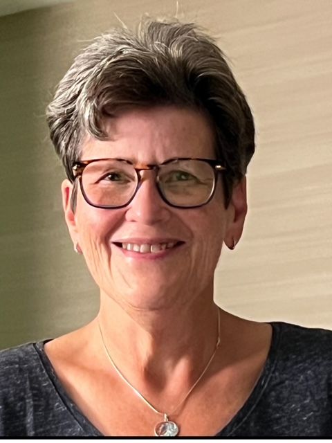 A white older woman with dark gray hair and round brown glasses