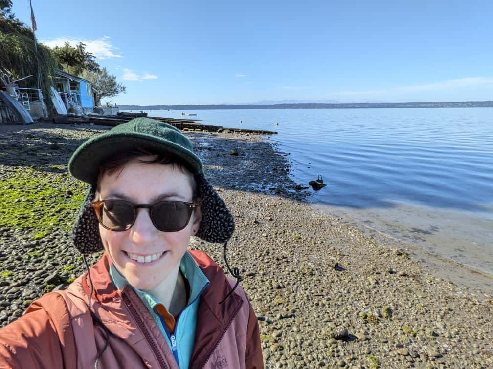 A white person wearing a floppy eared brim hat with dark sunglasses in front of some body of water