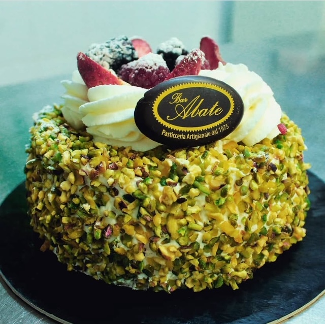 Una torta rotonda ricoperta di pistacchi tritati, guarnita con crema bianca, fragole fresche e una targhetta di cioccolato Bar Abate.