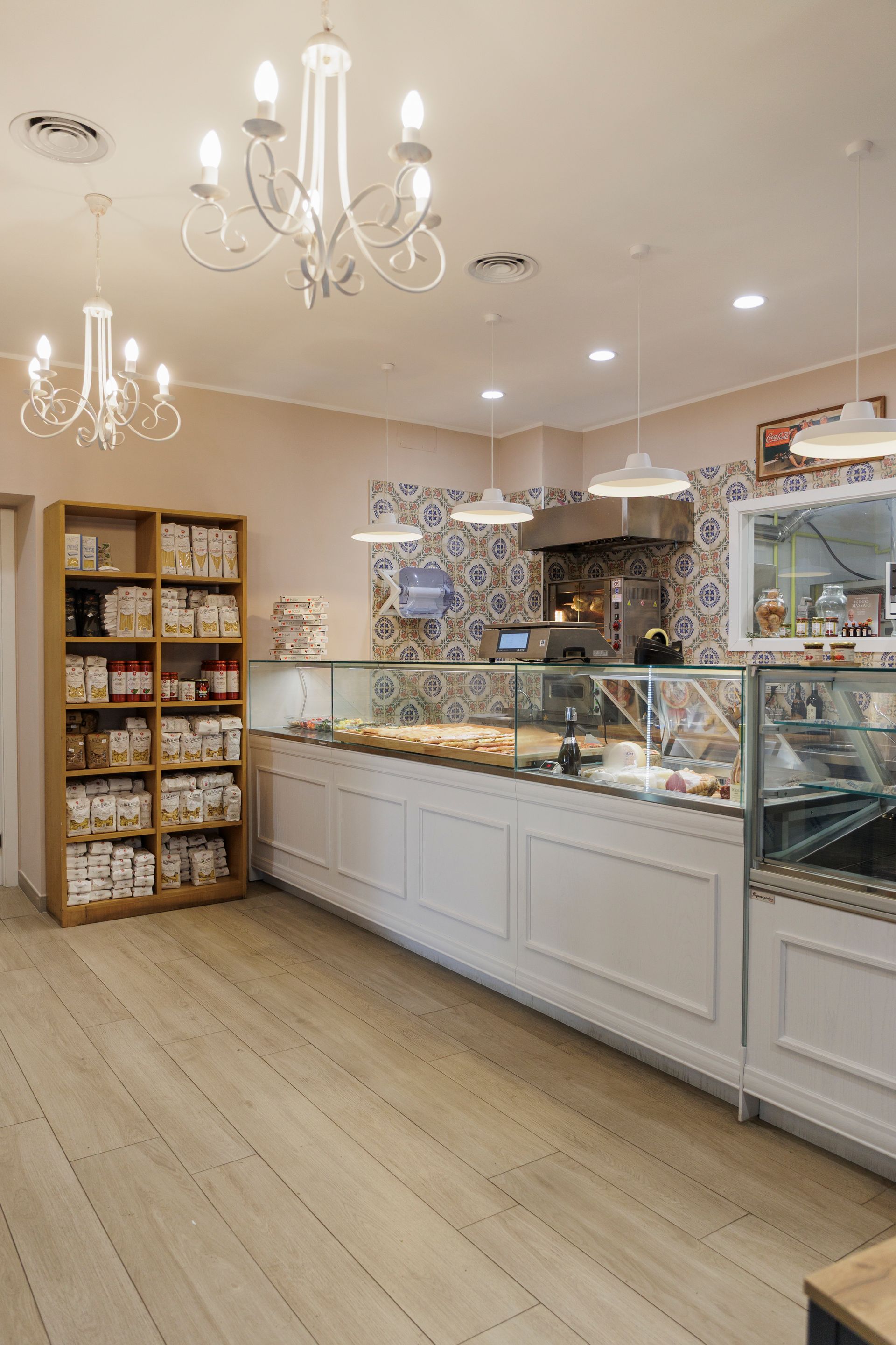 Un interno di panetteria pulito e luminoso, con mobili in legno bianco, un bancone espositivo in vetro, piastrelle decorate e lampadari.