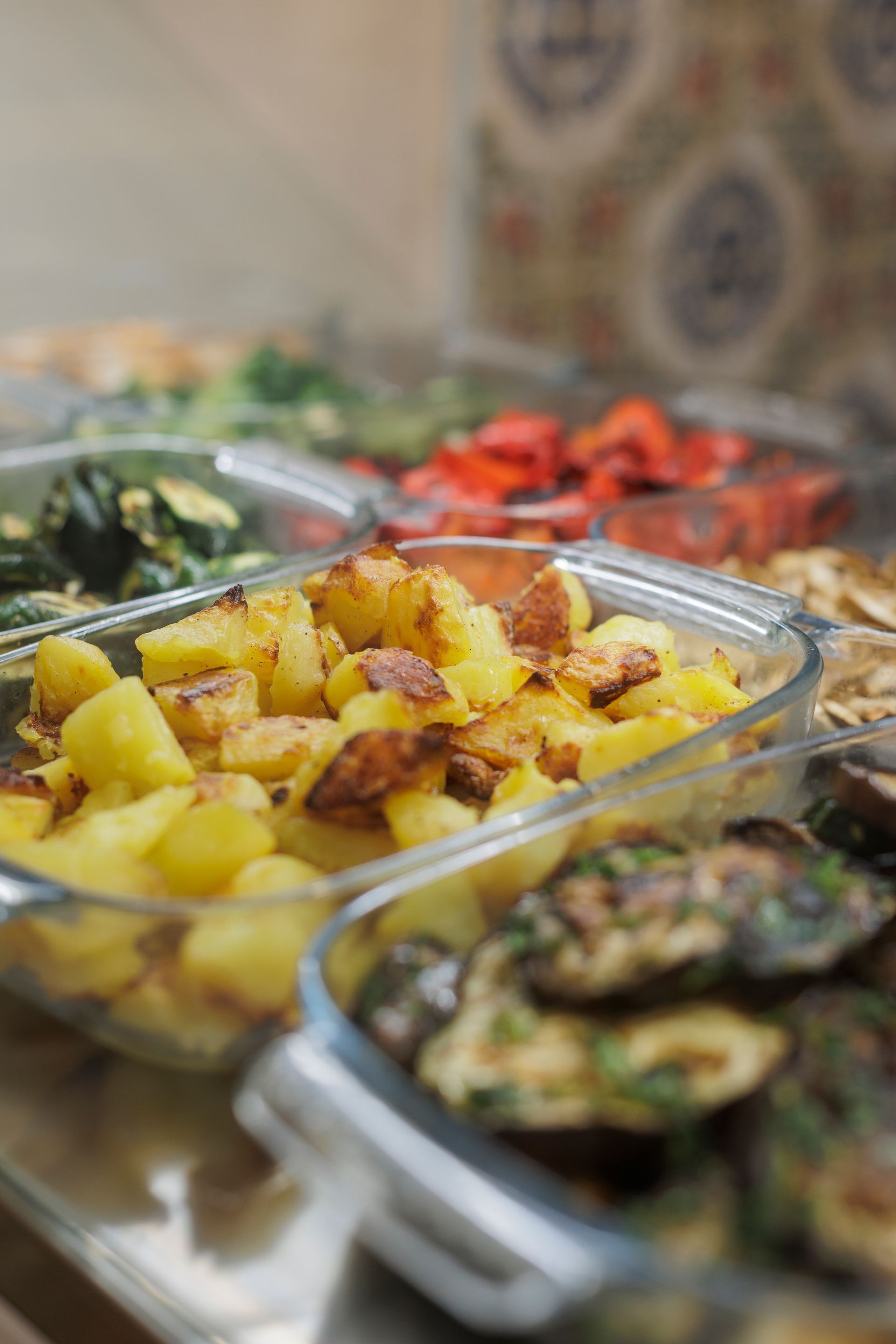 Piatti di vetro pieni di varie verdure arrostite, tra cui patate dorate, peperoni e zucchine, su un buffet.