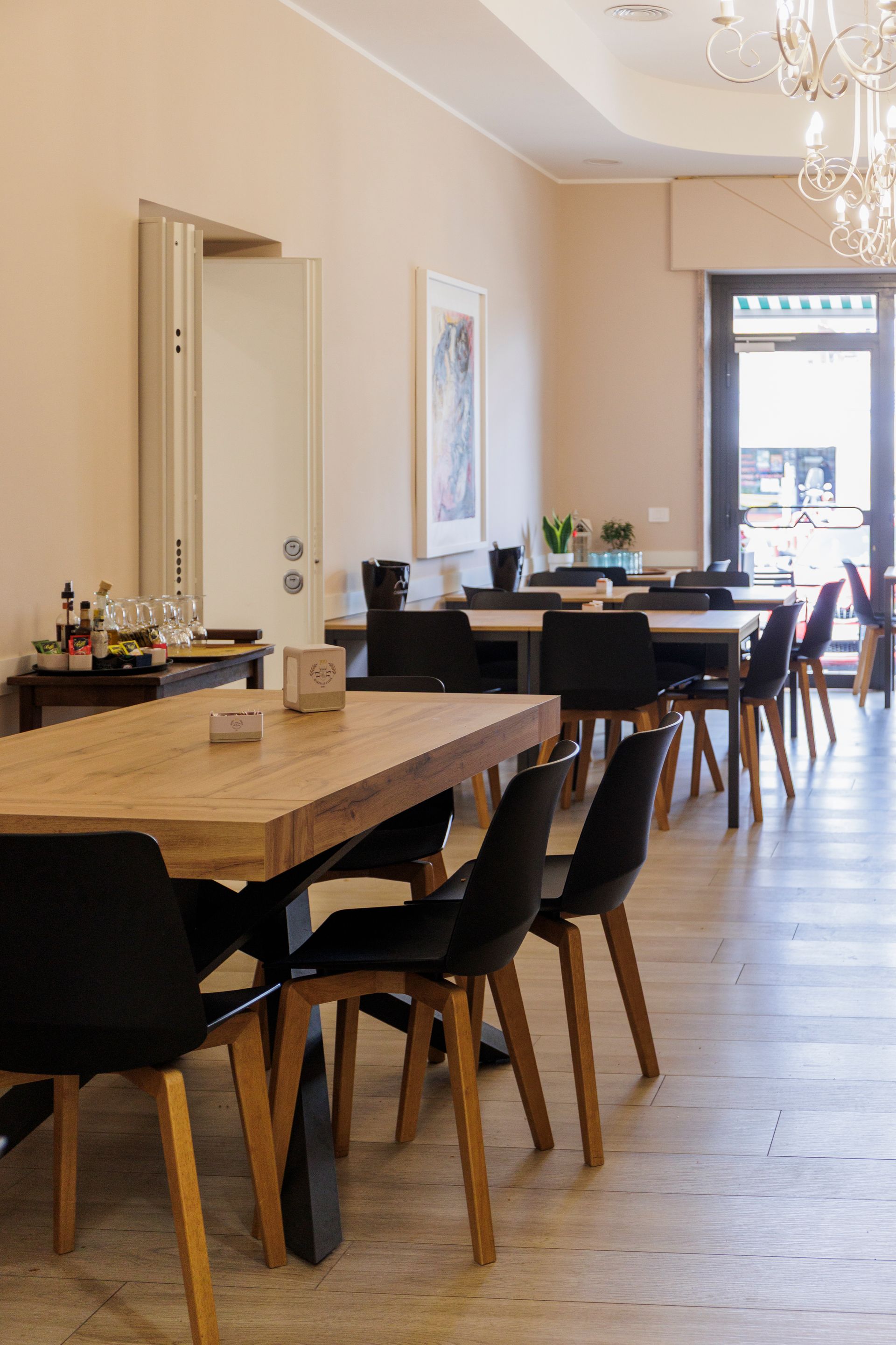 Una sala da pranzo moderna con tavoli in legno chiaro, sedie nere, pareti color pesca tenue e un elegante lampadario di cristallo.