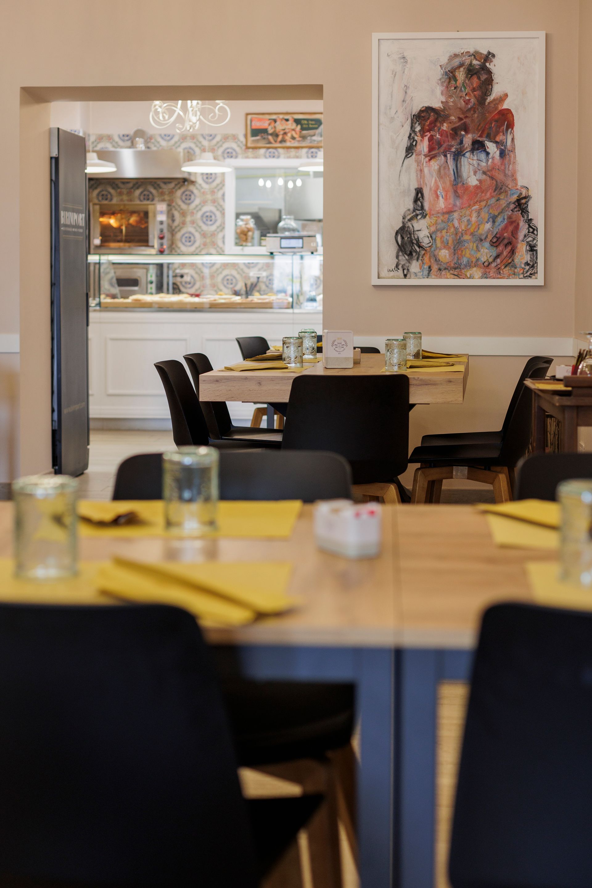 Interno di un ristorante con tavoli apparecchiati con tovagliette gialle, sedie nere e un quadro su una parete chiara.