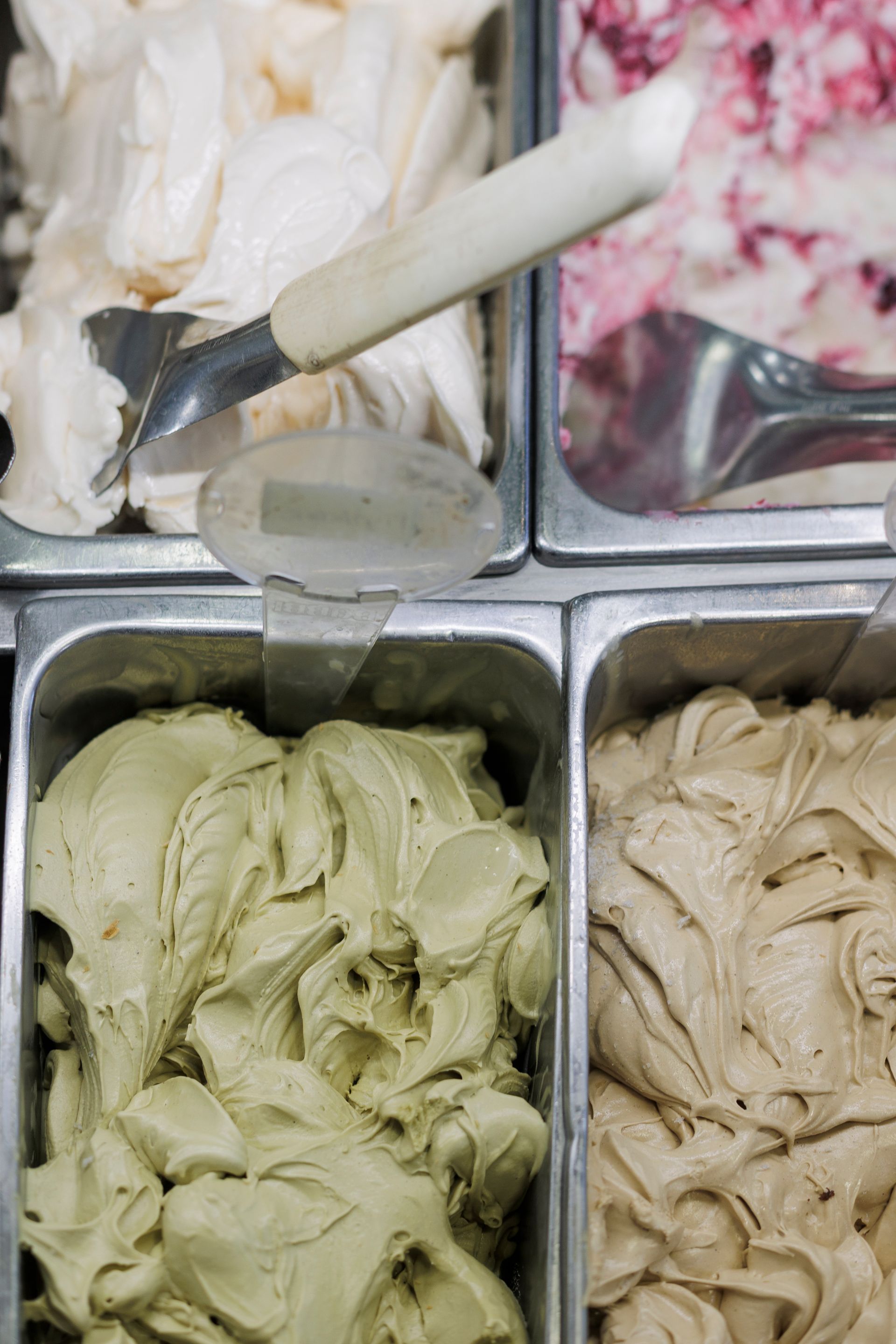 Quattro vaschette di metallo riempite con gelato o sorbetto di vari colori, con un cucchiaio da portata argentato appoggiato in uno dei contenitori.
