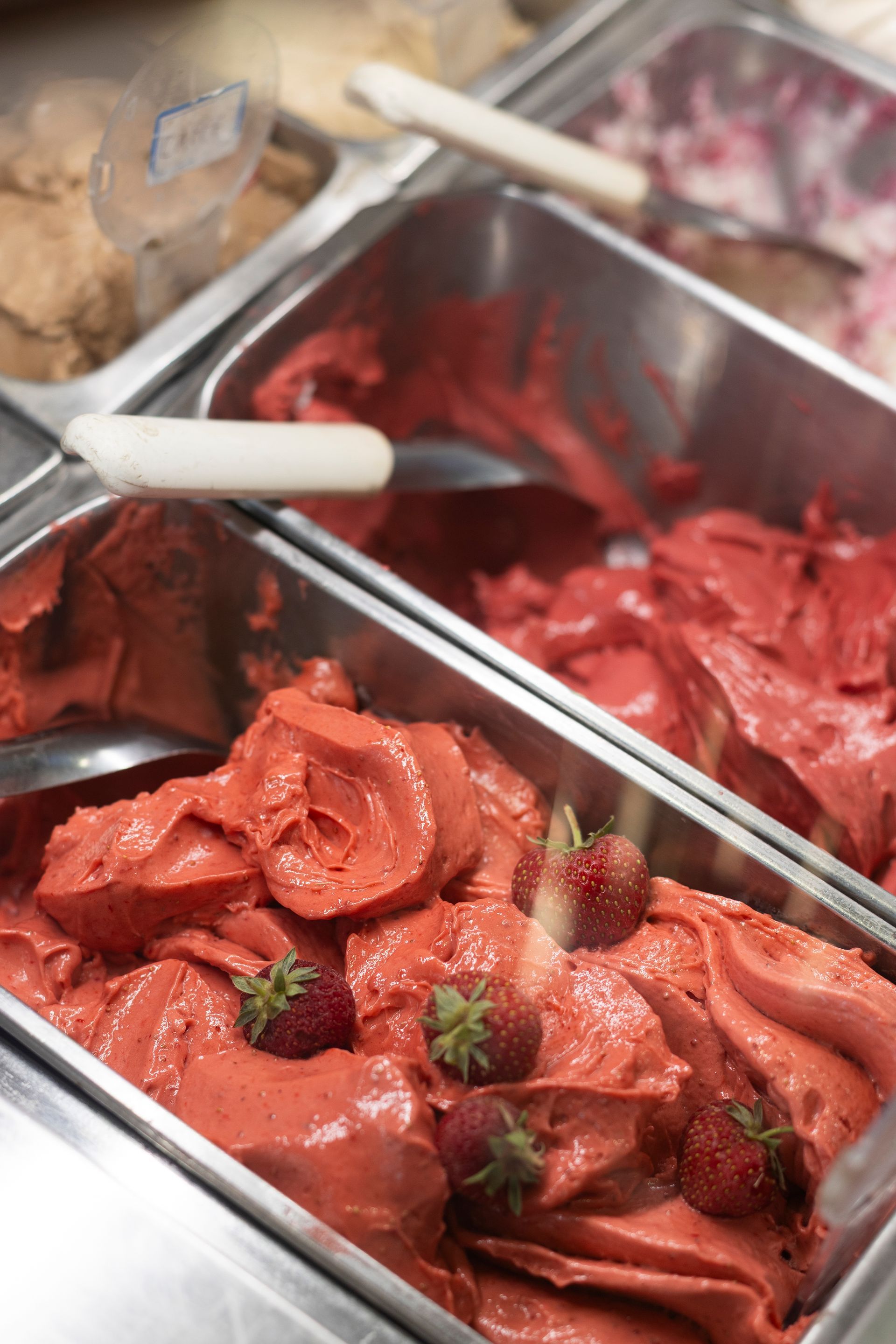 Contenitori di metallo pieni di palline di gelato alla fragola rossa guarnite con fragole fresche intere, con altri gusti disponibili nelle vicinanze.