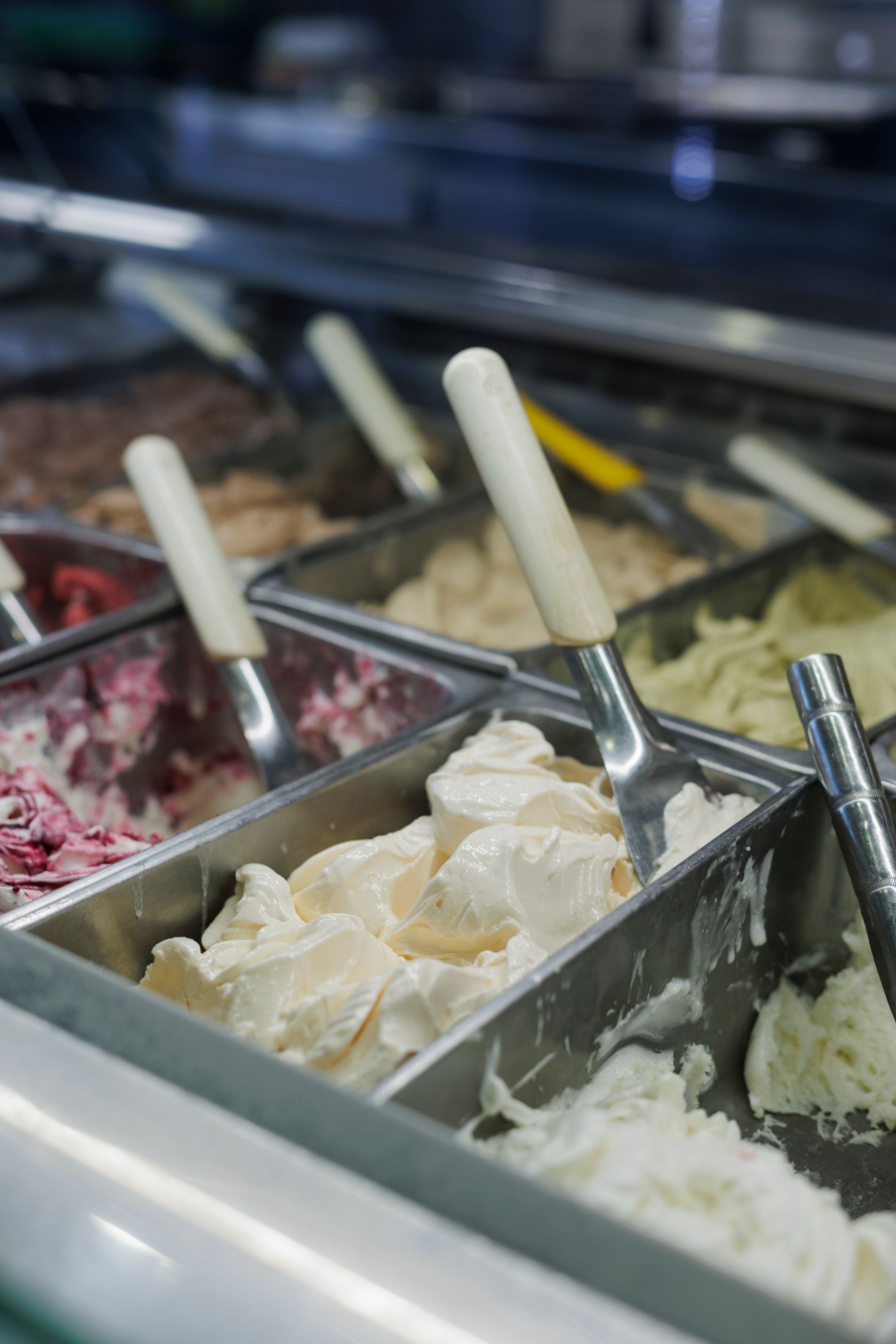 Un primo piano di gelato in vaschette di acciaio inox all'interno di una vetrina, ciascuna con una paletta per servirlo.