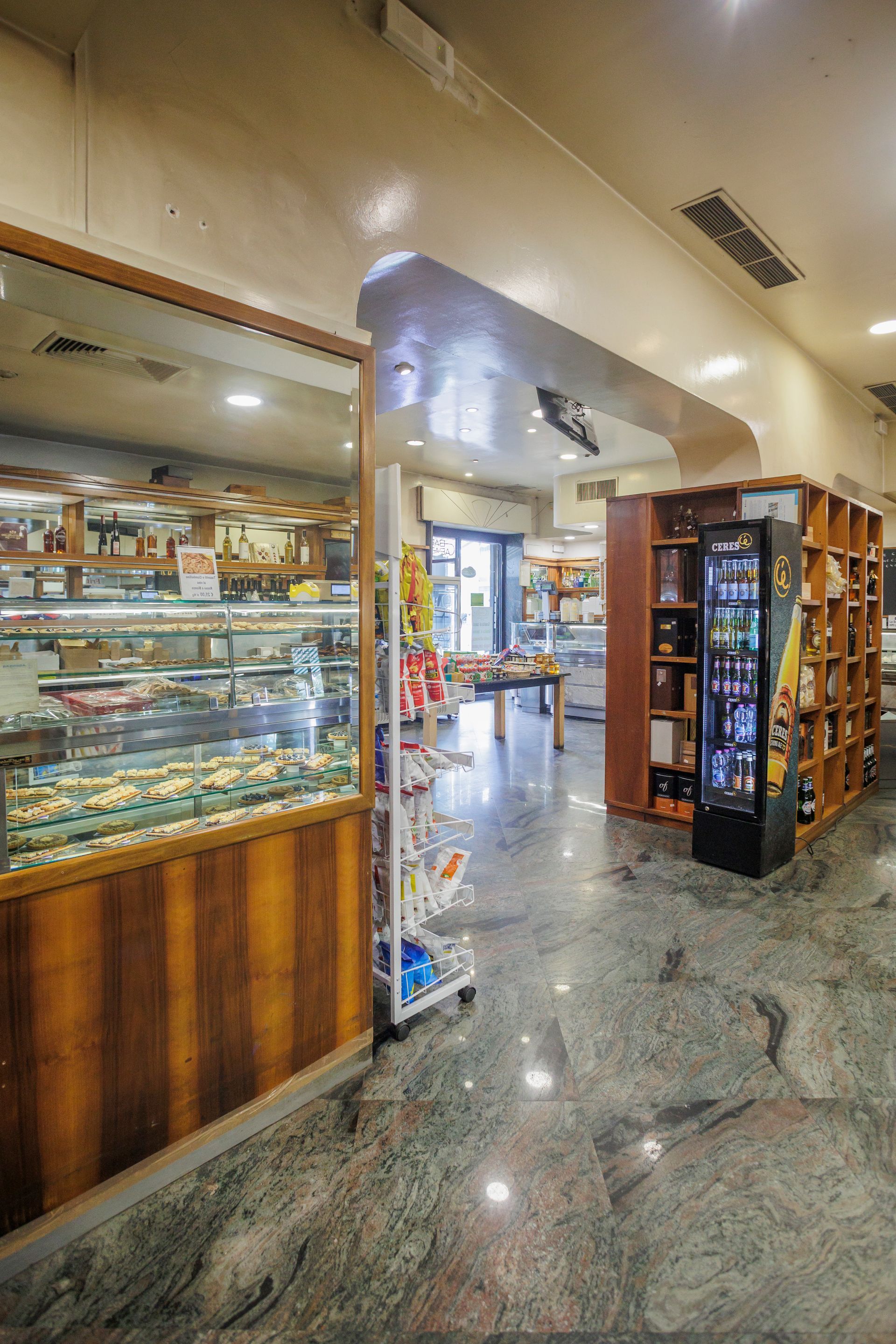 Vista interna di un mercato ben illuminato con vetrina, scaffali in legno, distributore automatico e pavimenti in marmo.