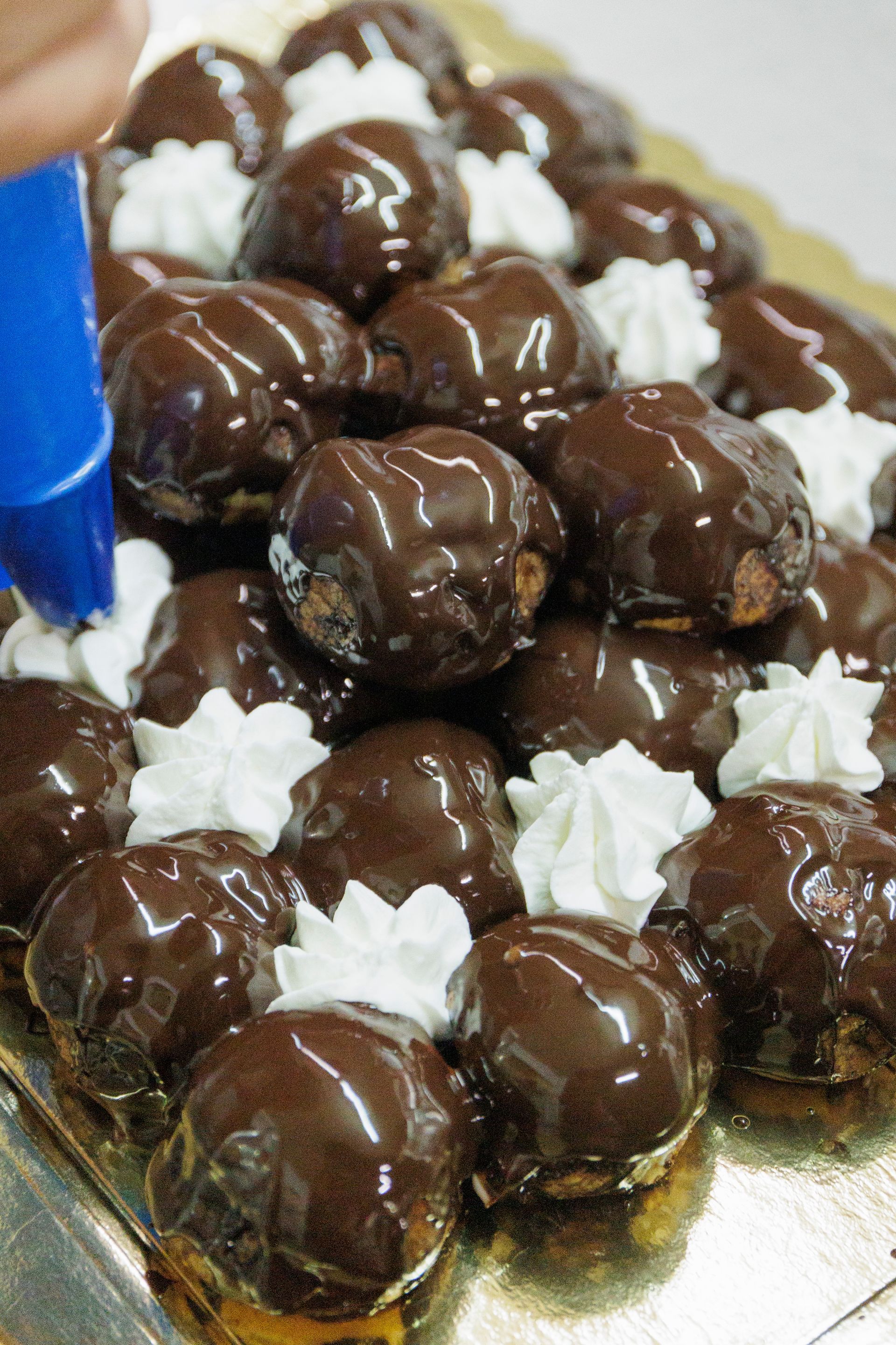 Una persona usa una sac à poche blu per decorare un vassoio di bignè ricoperti di glassa al cioccolato con riccioli di crema bianca.