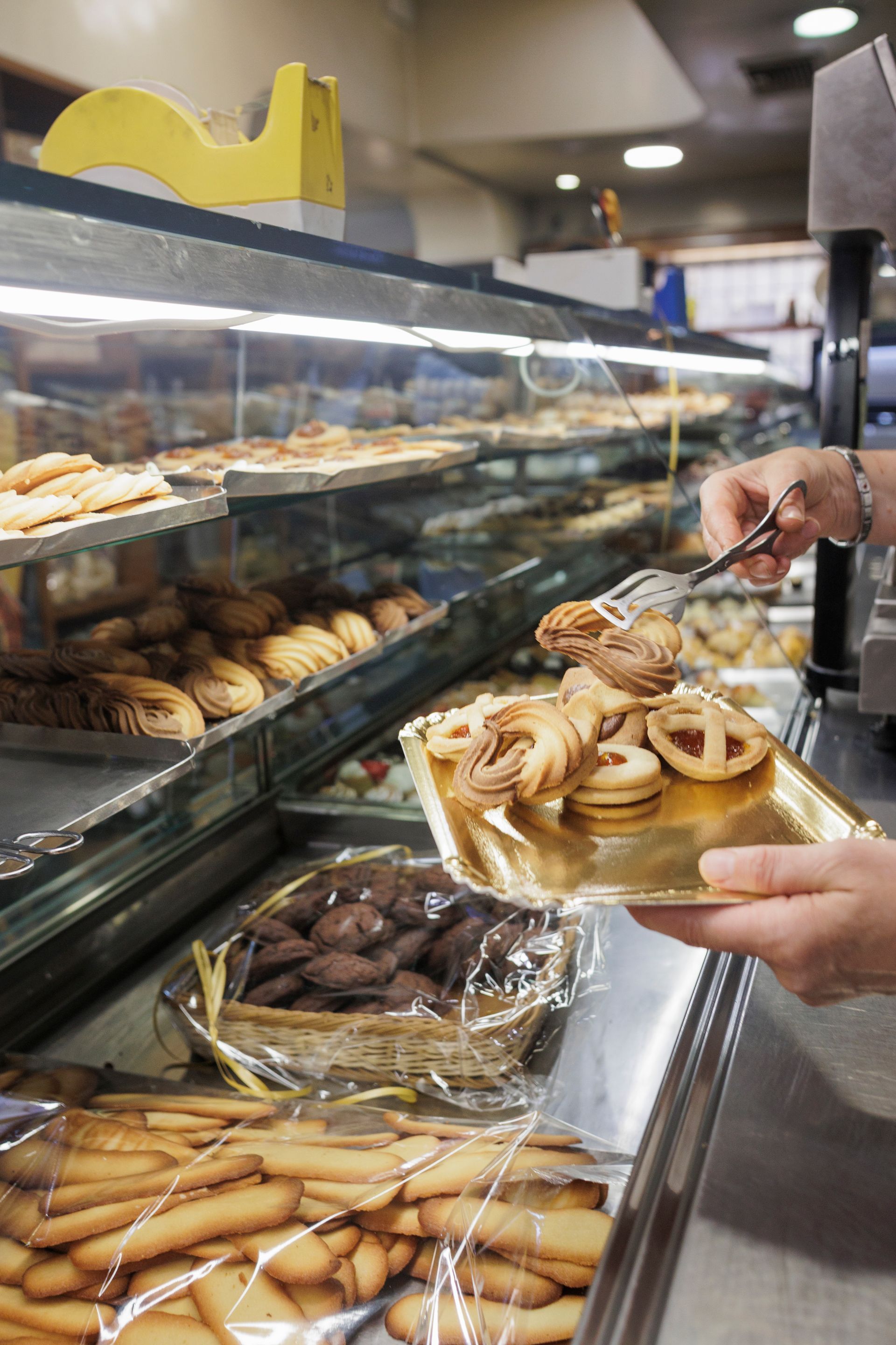 Una persona usa delle pinze per disporre vari biscotti su un vassoio dorato in una vetrina di una pasticceria piena di pasticcini assortiti.
