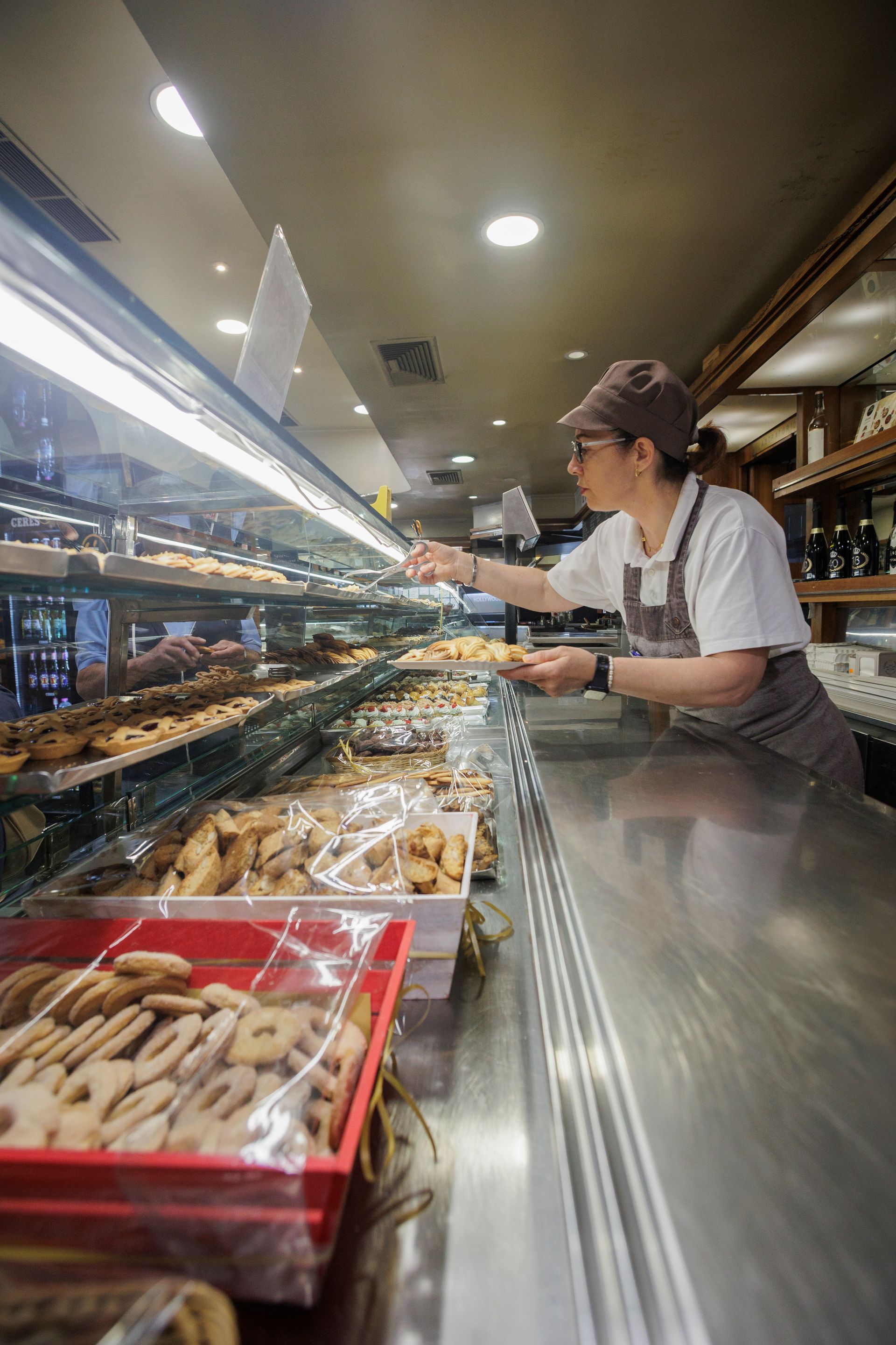 Una persona con un cappello e un grembiule marroni lavora dietro il bancone di una pasticceria, sistemando vassoi di biscotti all'interno di una vetrina.