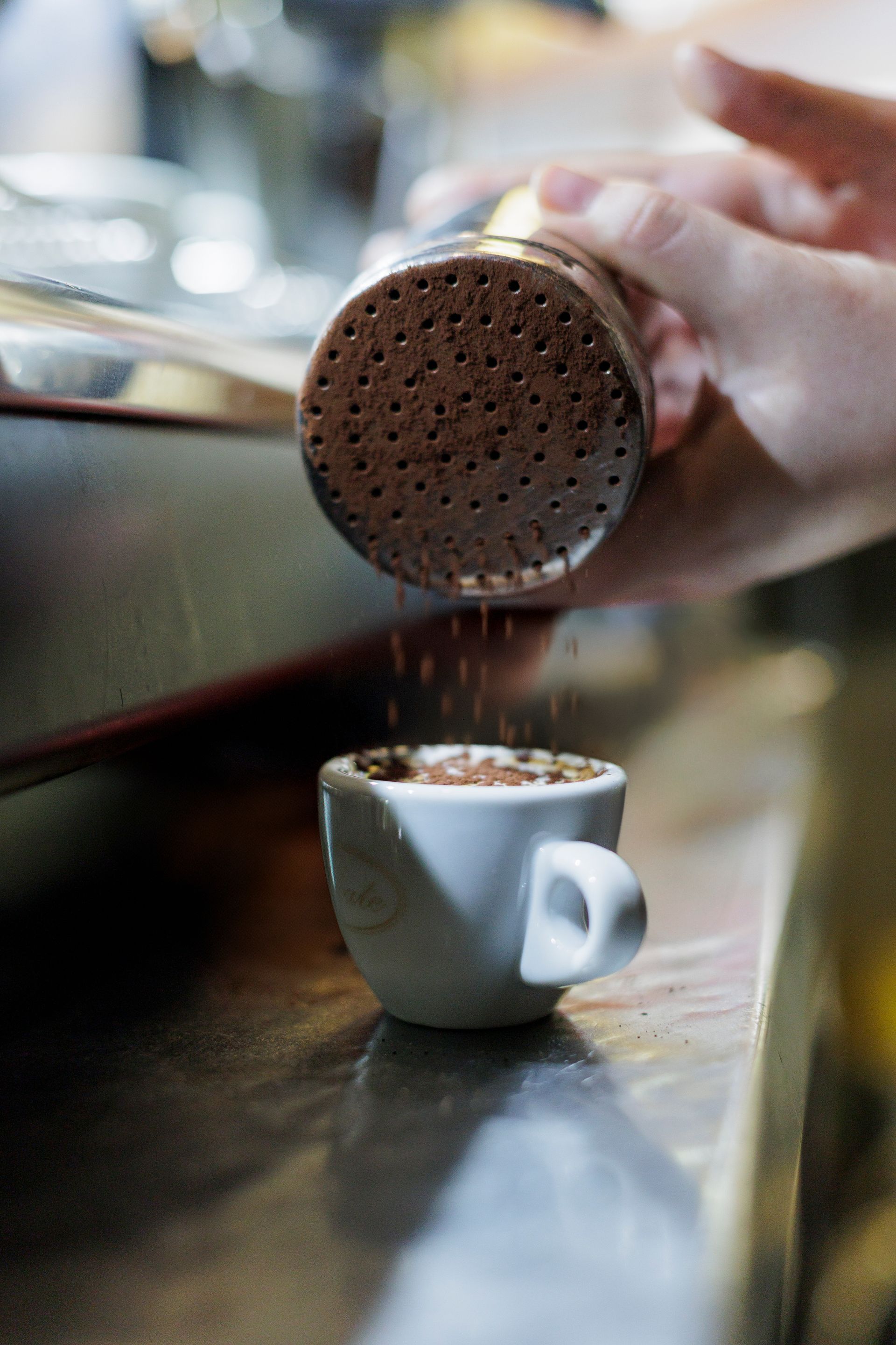 Una mano sparge cacao in polvere da uno spargitore su una piccola tazza bianca di caffè espresso appoggiata su un bancone.
